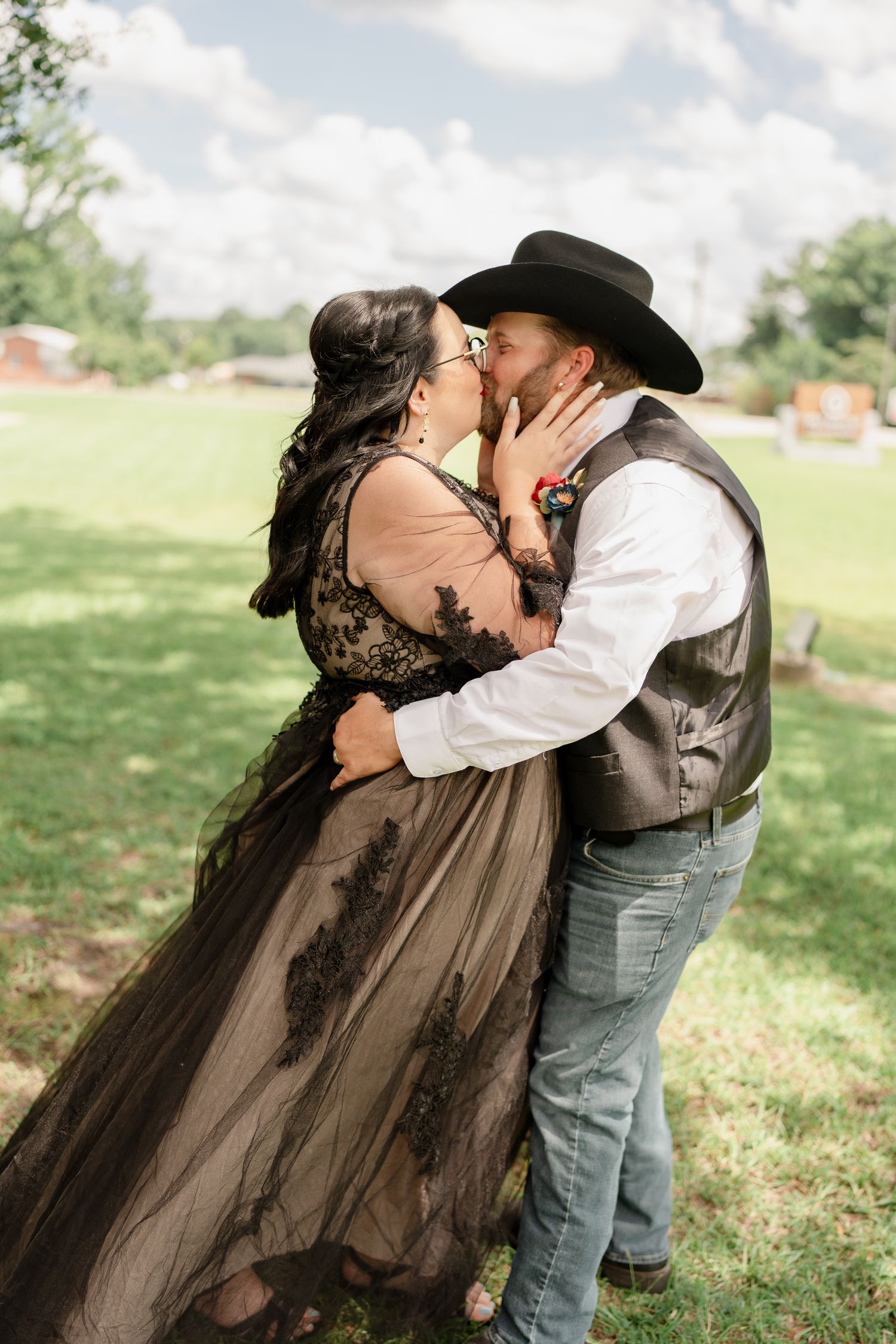 A romantic couple embraces and kisses outdoors, with her wearing a black lace dress and him in cowboy attire.