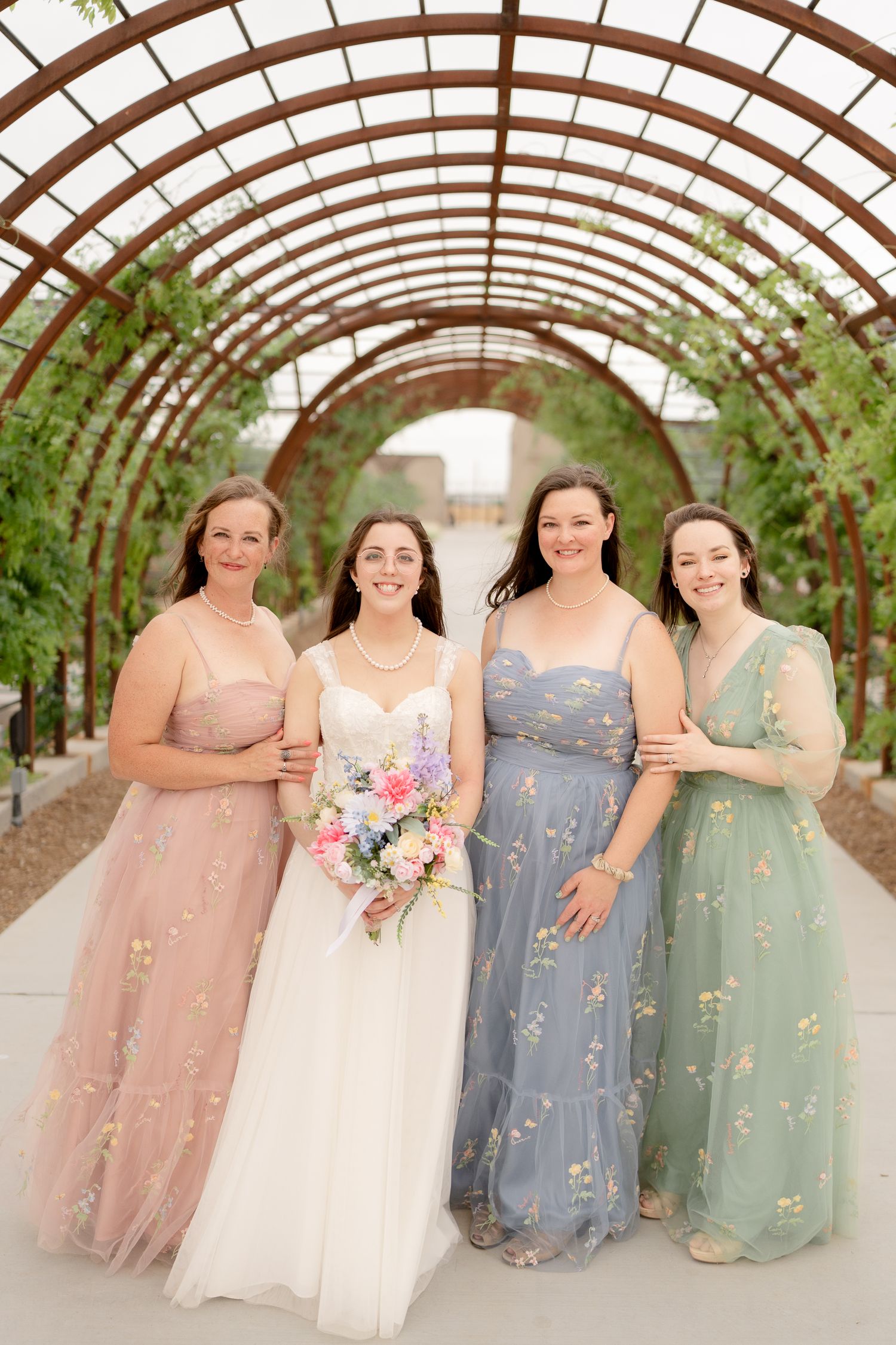 A bridal party wearing pastel dresses poses together under a curved greenhouse archway with climbing vines.