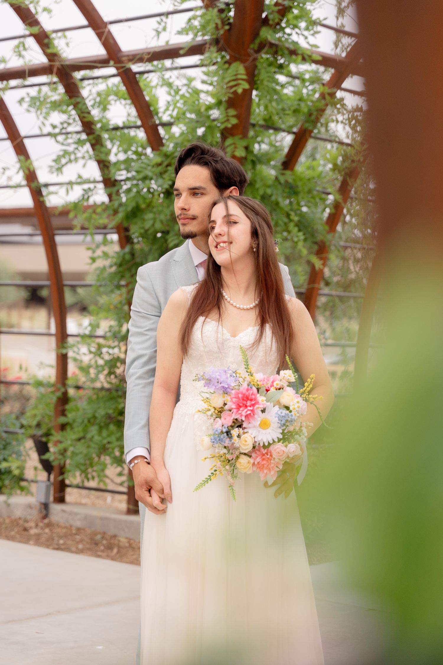 Wedding couple poses with colorful bouquet in a greenhouse corridor lined with climbing plants.