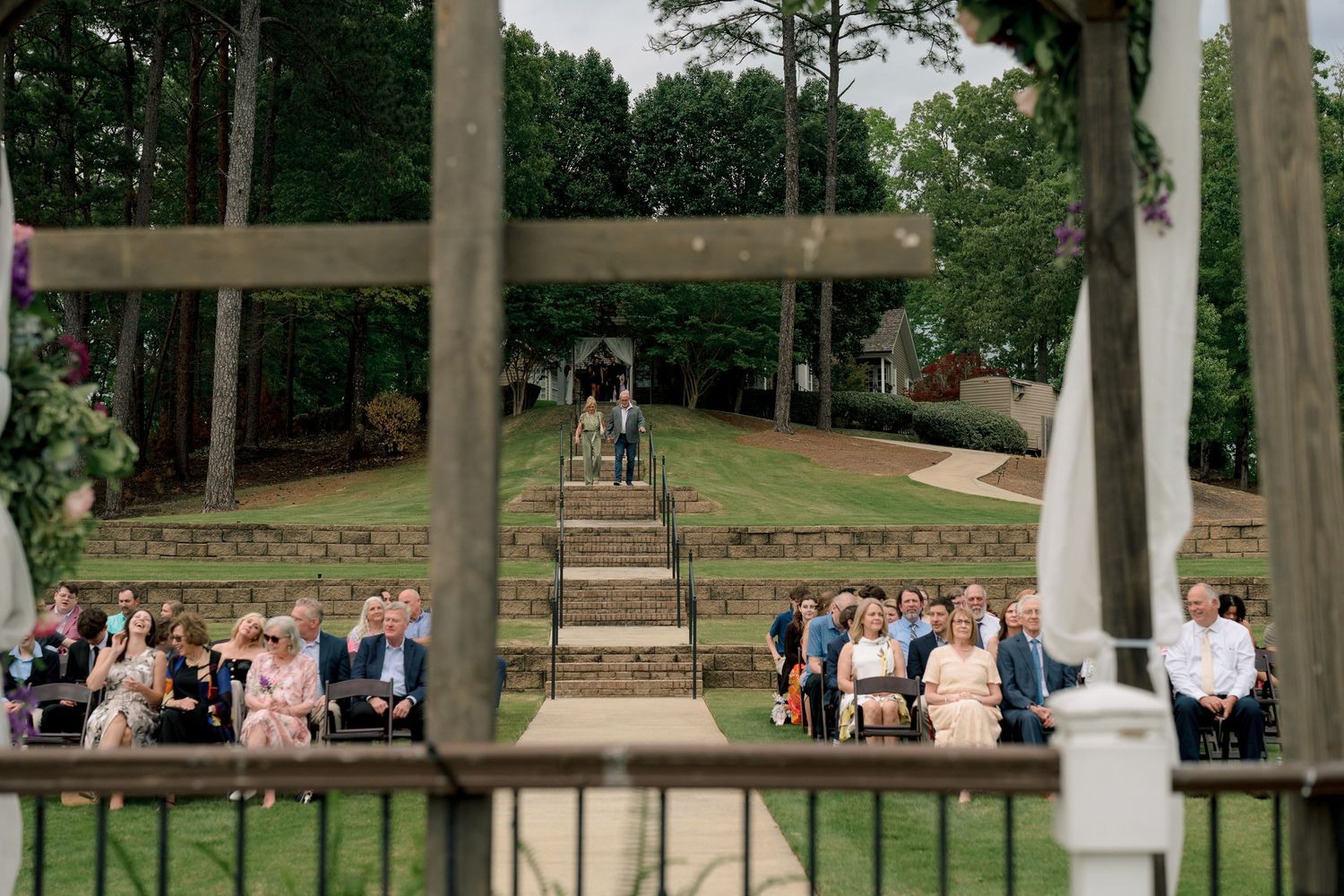 Outdoor wedding ceremony with guests seated on tiered stone steps surrounded by lush green trees.