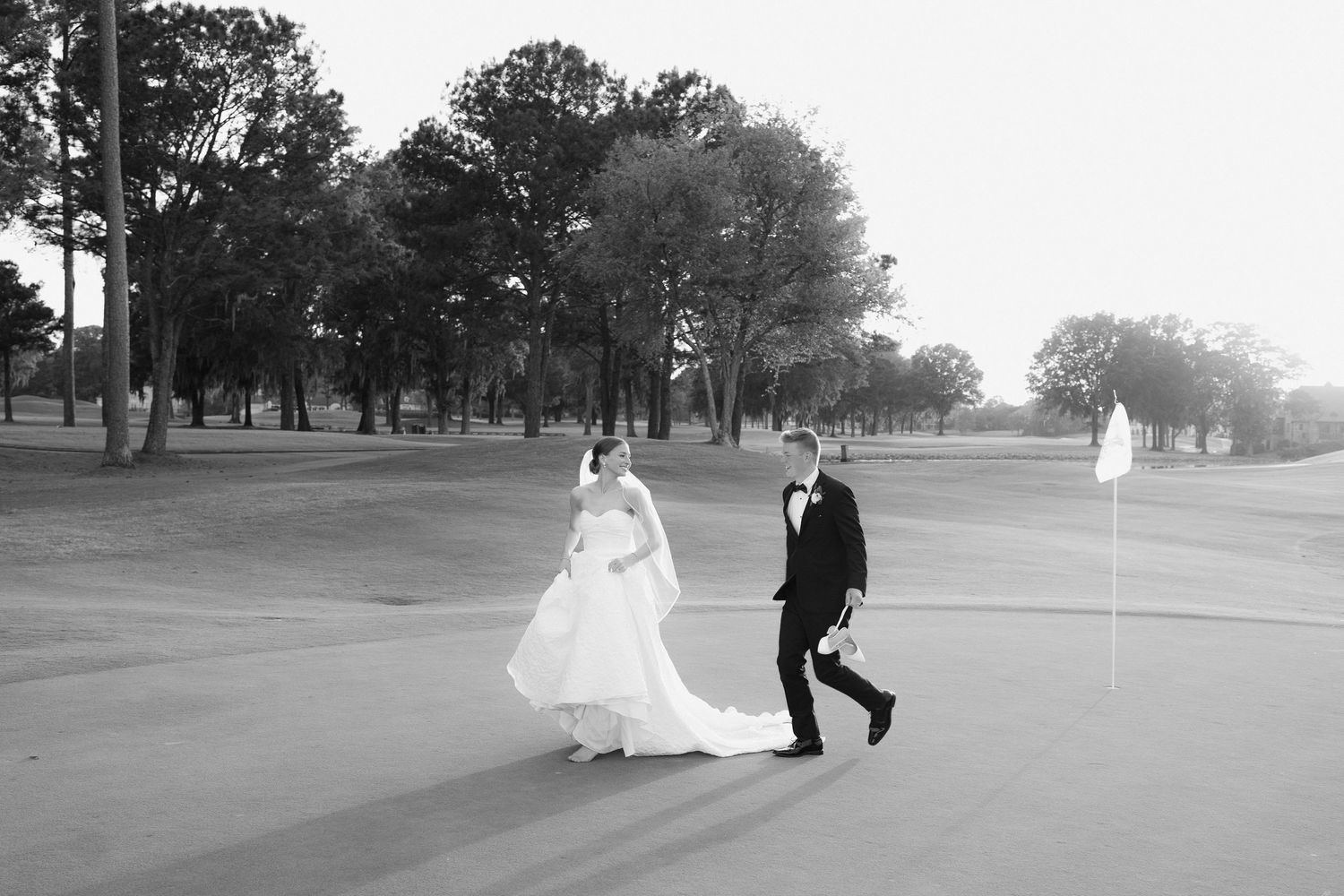Black and white photo of a bride and groom walking on a golf course in wedding attire.