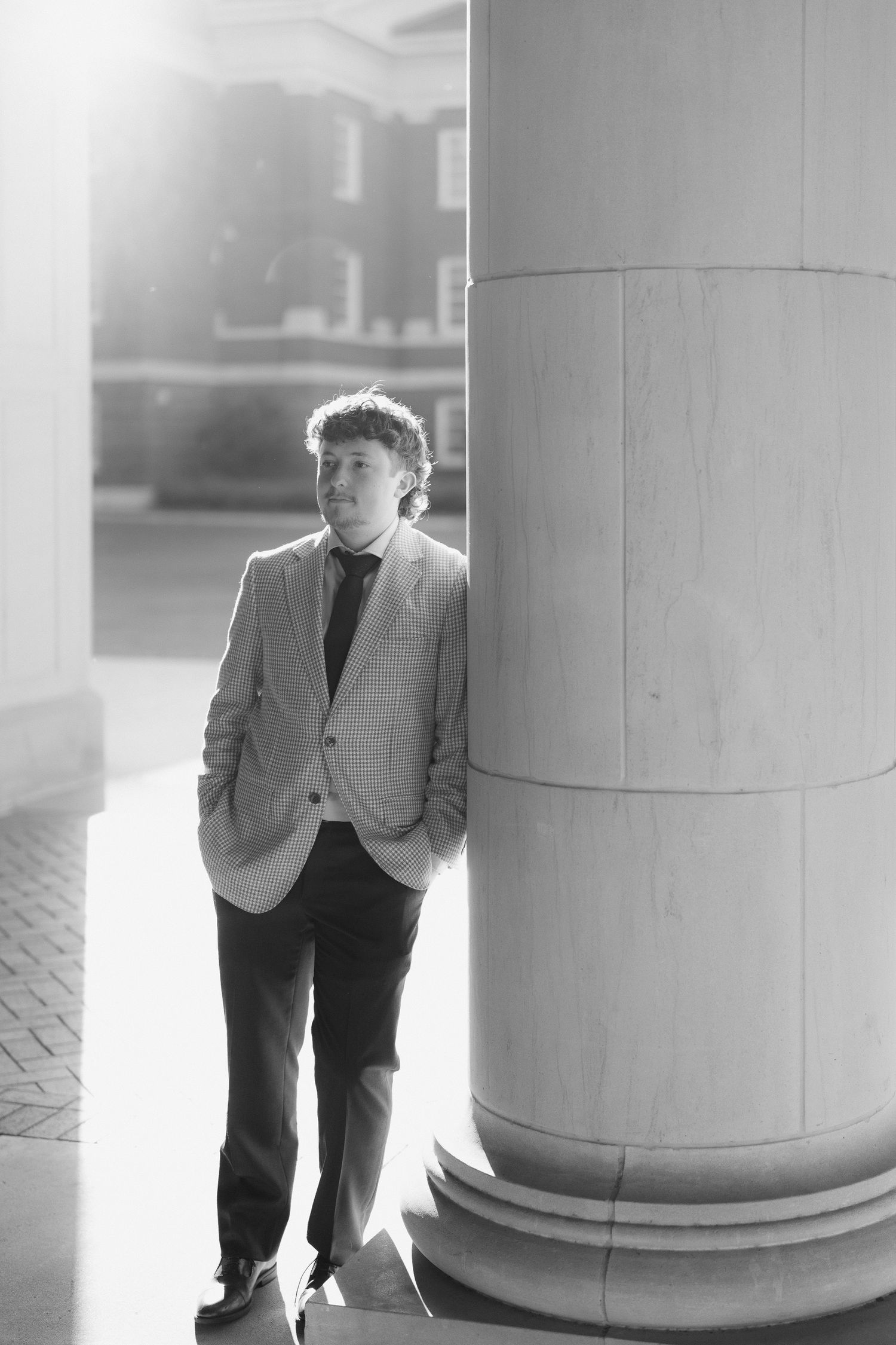 Black and white portrait of a young man leaning against a large column on a college campus.