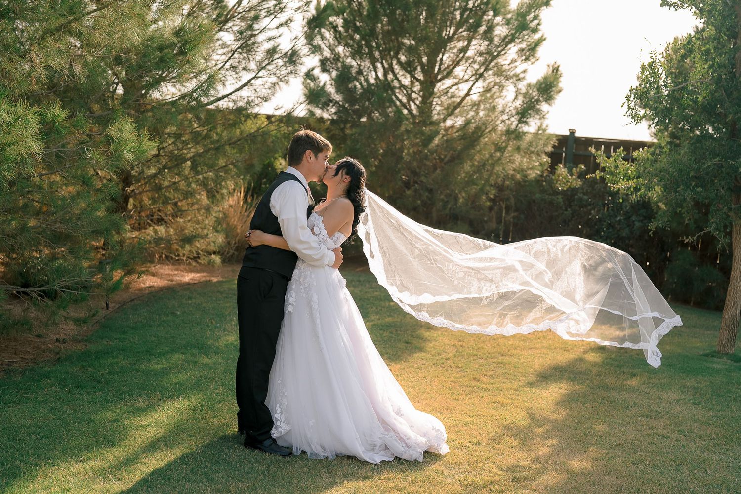 Bride and groom kissing outdoors with wedding veil dramatically flowing in the wind at golden hour.