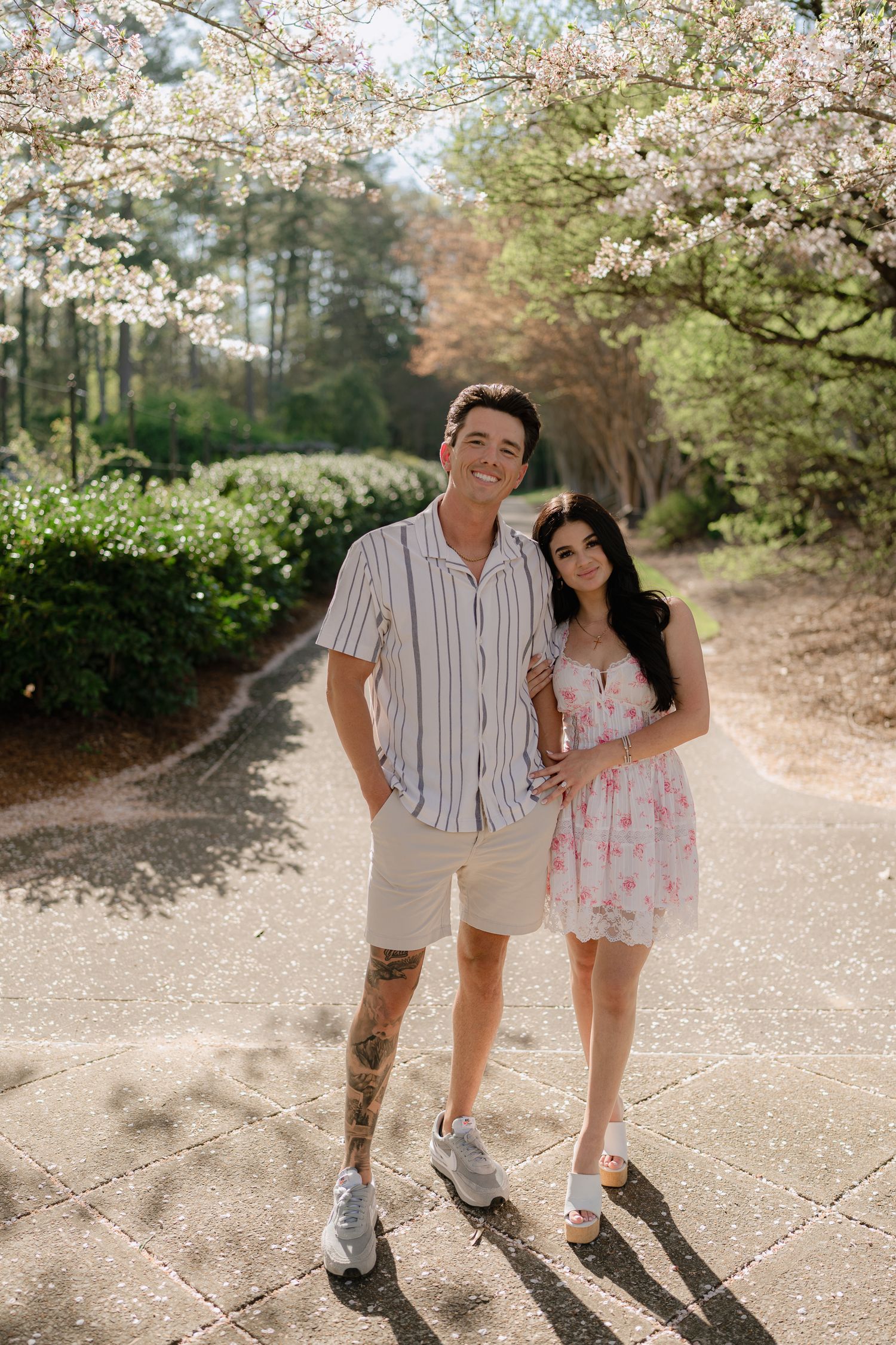 Smiling couple poses together under blooming cherry blossom trees on a bright spring day in a scenic garden.