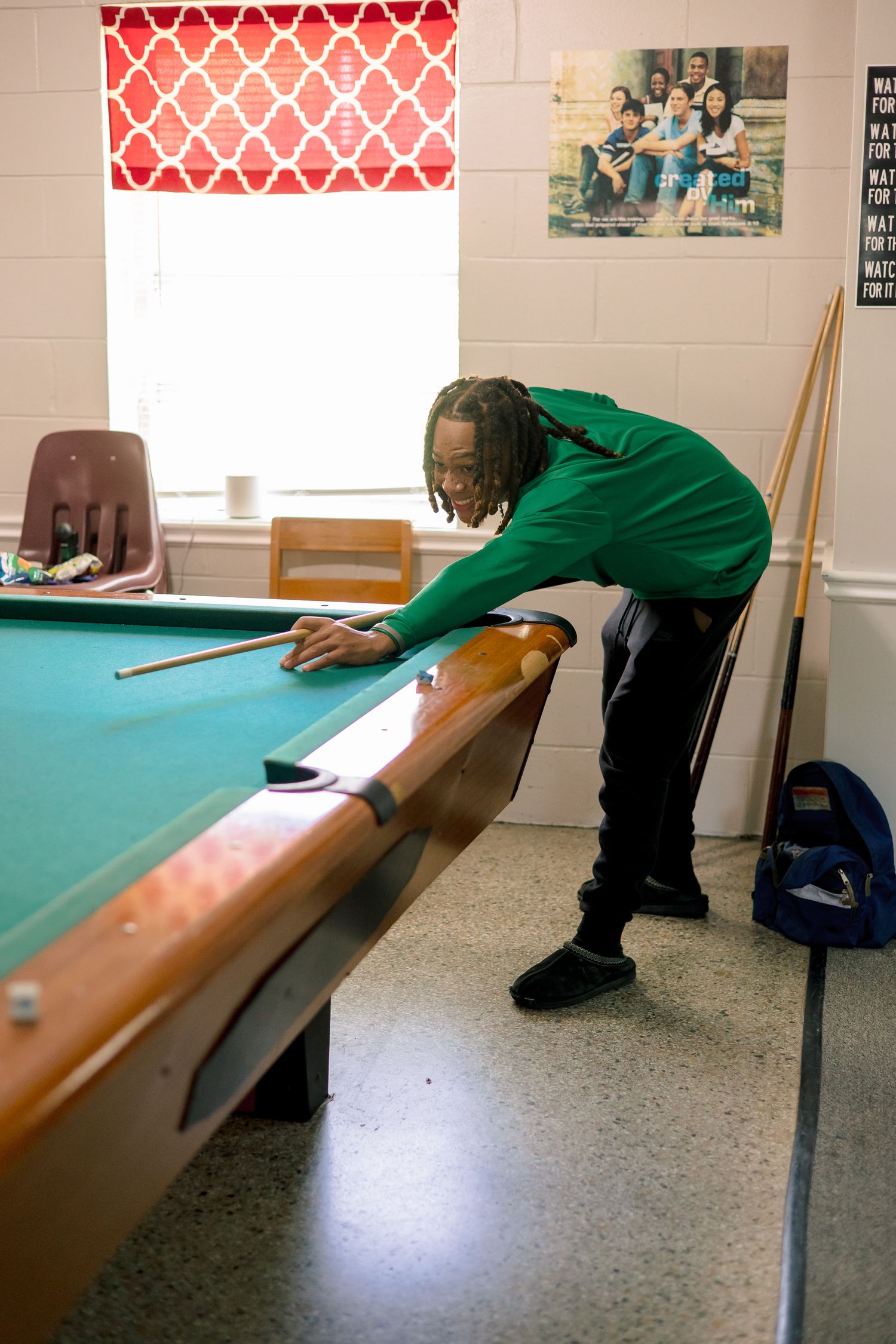Man in green shirt leaning over pool table preparing to take a shot with a cue stick.