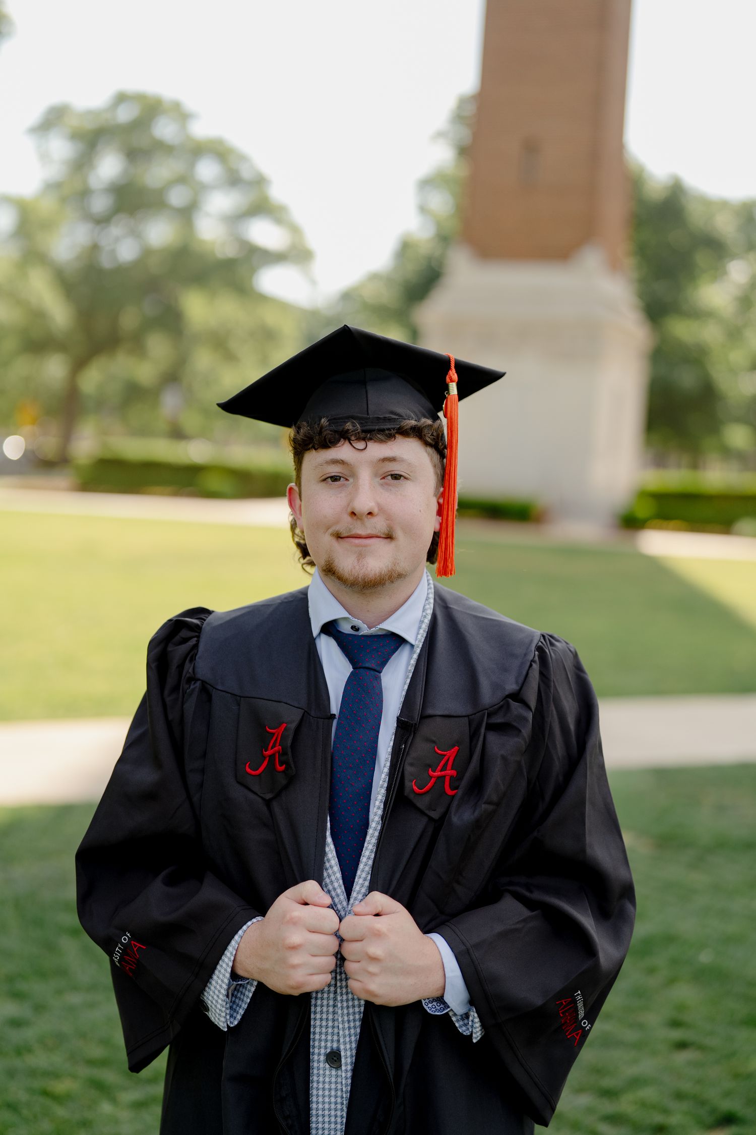 Young man in University of Alabama graduation cap and gown smiling outdoors on campus.