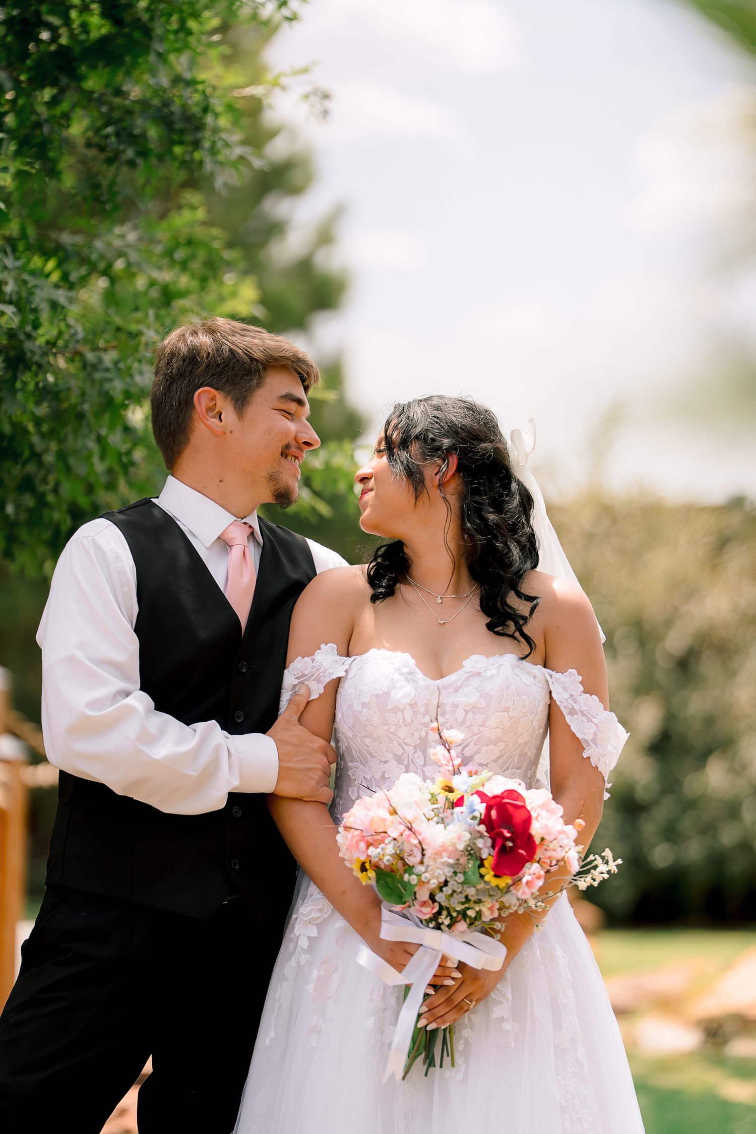 Bride and groom share a romantic moment outdoors, bride holding a pink and red bouquet in a lace off-shoulder wedding gown.