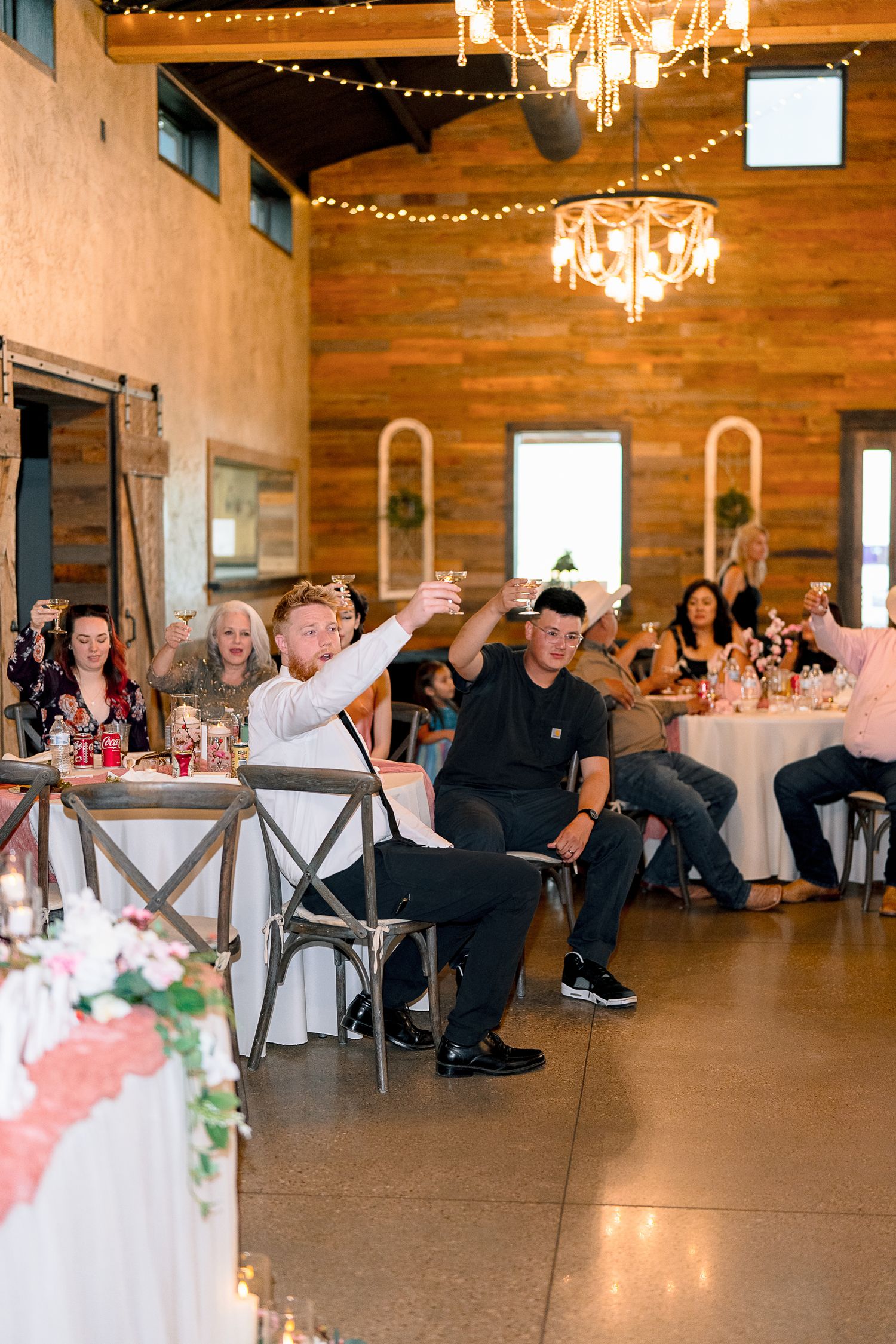 Wedding guests raise glasses in toast during reception in rustic barn venue with string lights.