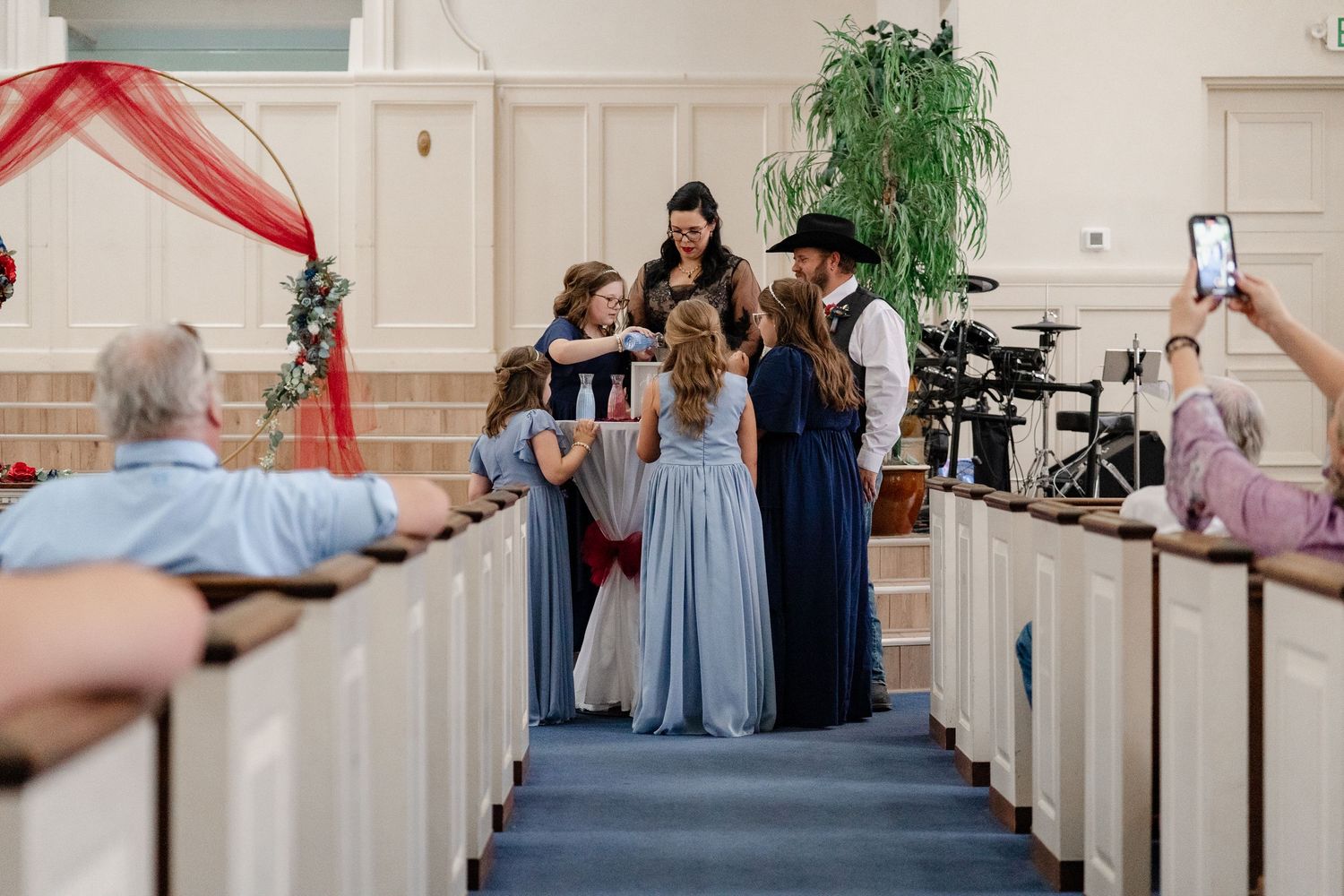 Church pews line a wedding ceremony aisle decorated with red tulle and greenery.