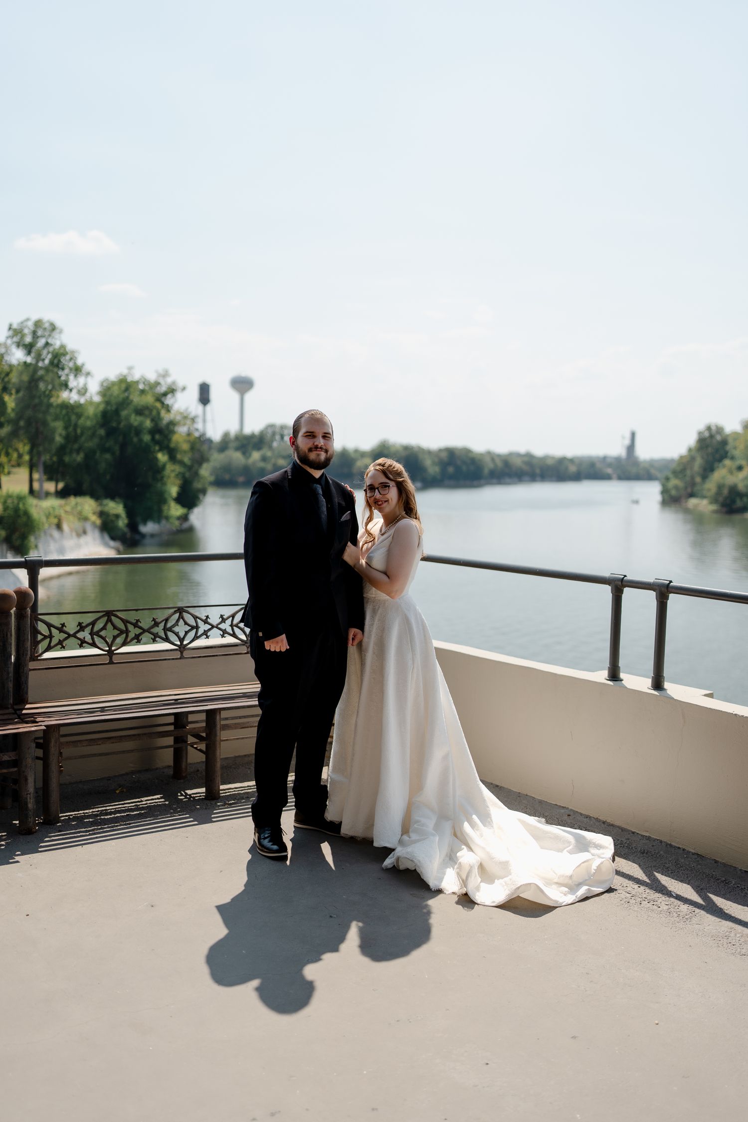 A couple stands on a waterfront balcony during their wedding day with a scenic lake view in the background.