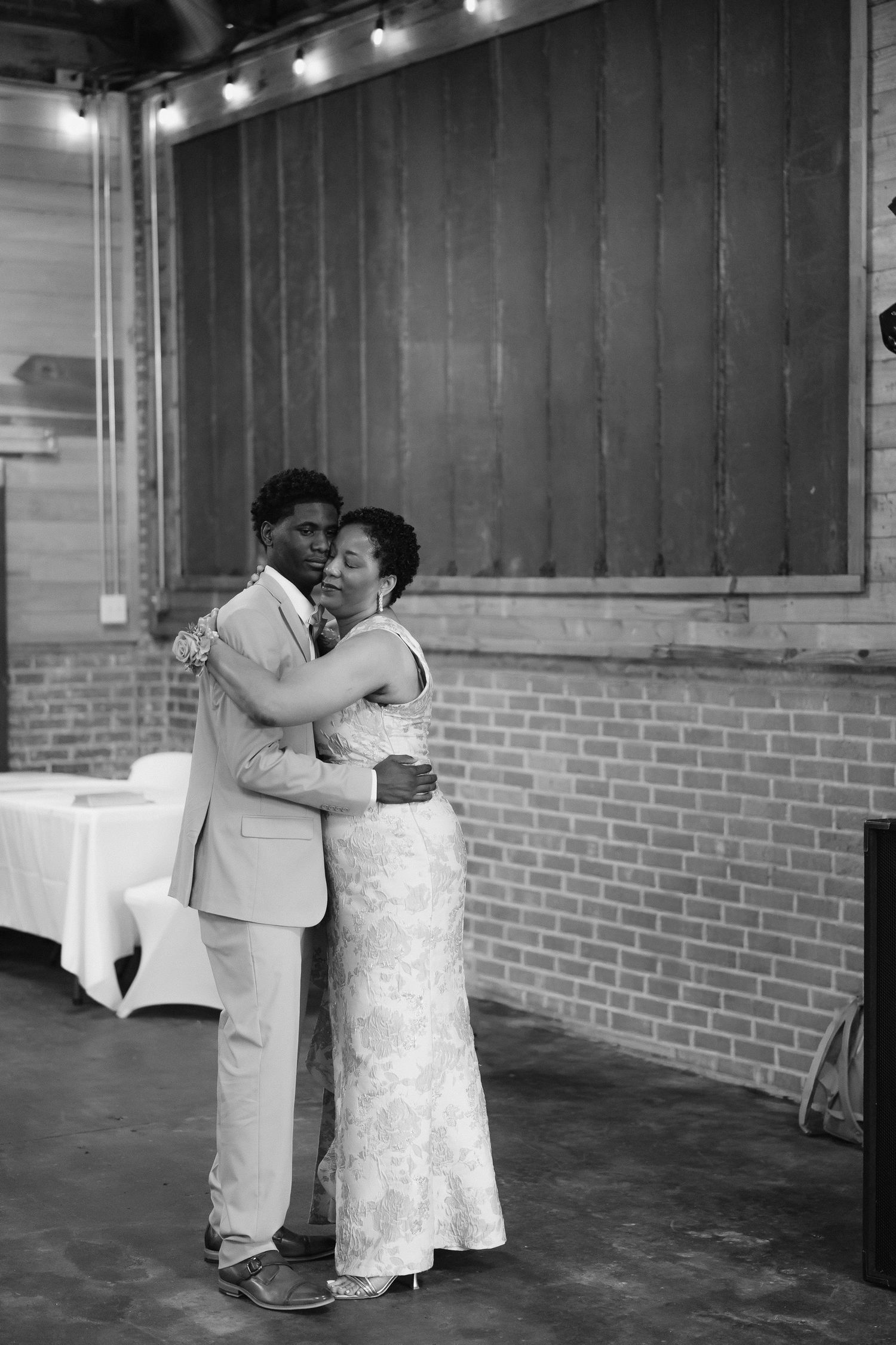 Black and white photo of groom in tan suit sharing emotional mother-son dance in rustic brick reception venue.