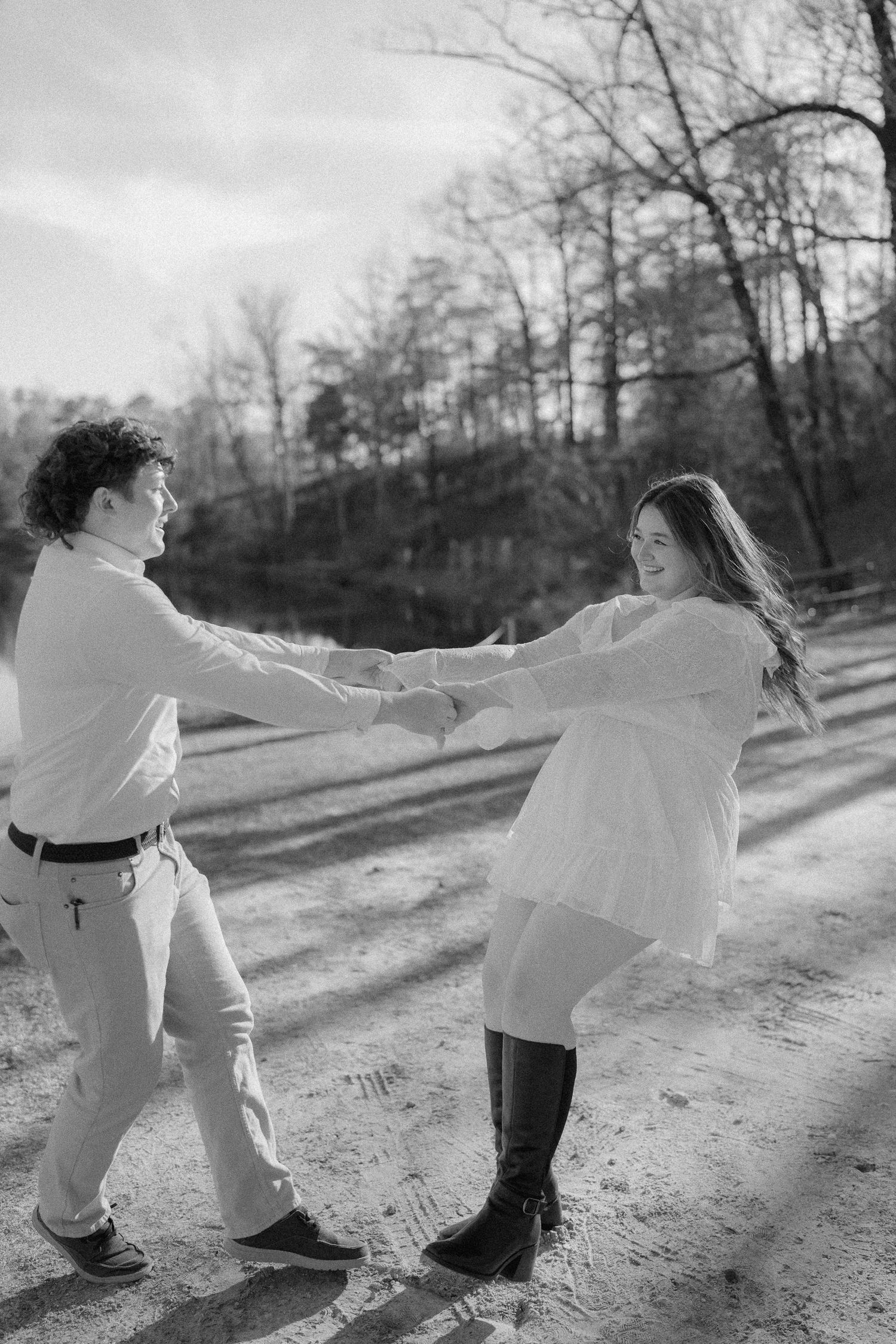 Black and white photo of couple holding hands and spinning playfully on a sandy outdoor path.