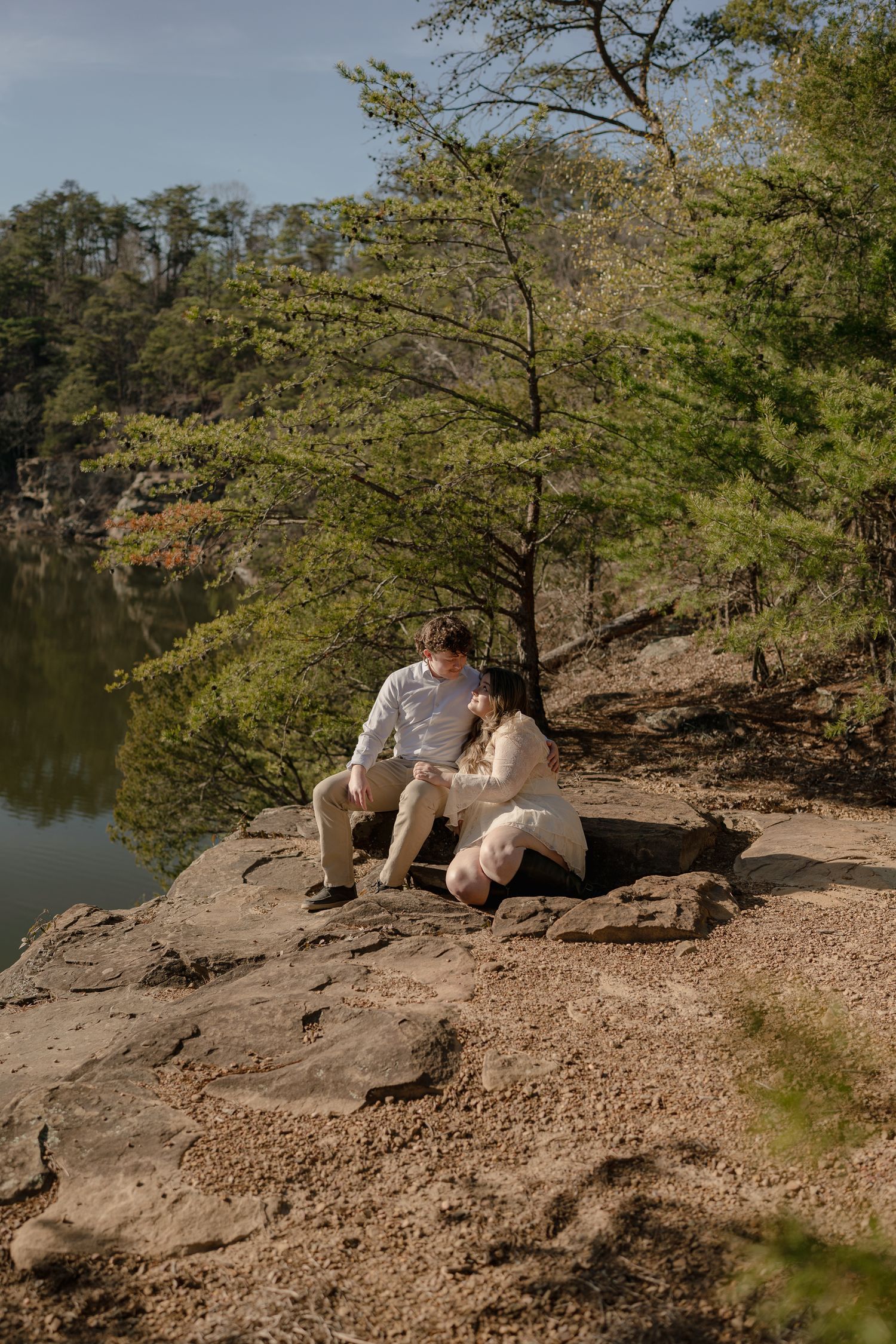 Couple sitting together on rocky ledge overlooking serene lake surrounded by lush green trees on sunny day.