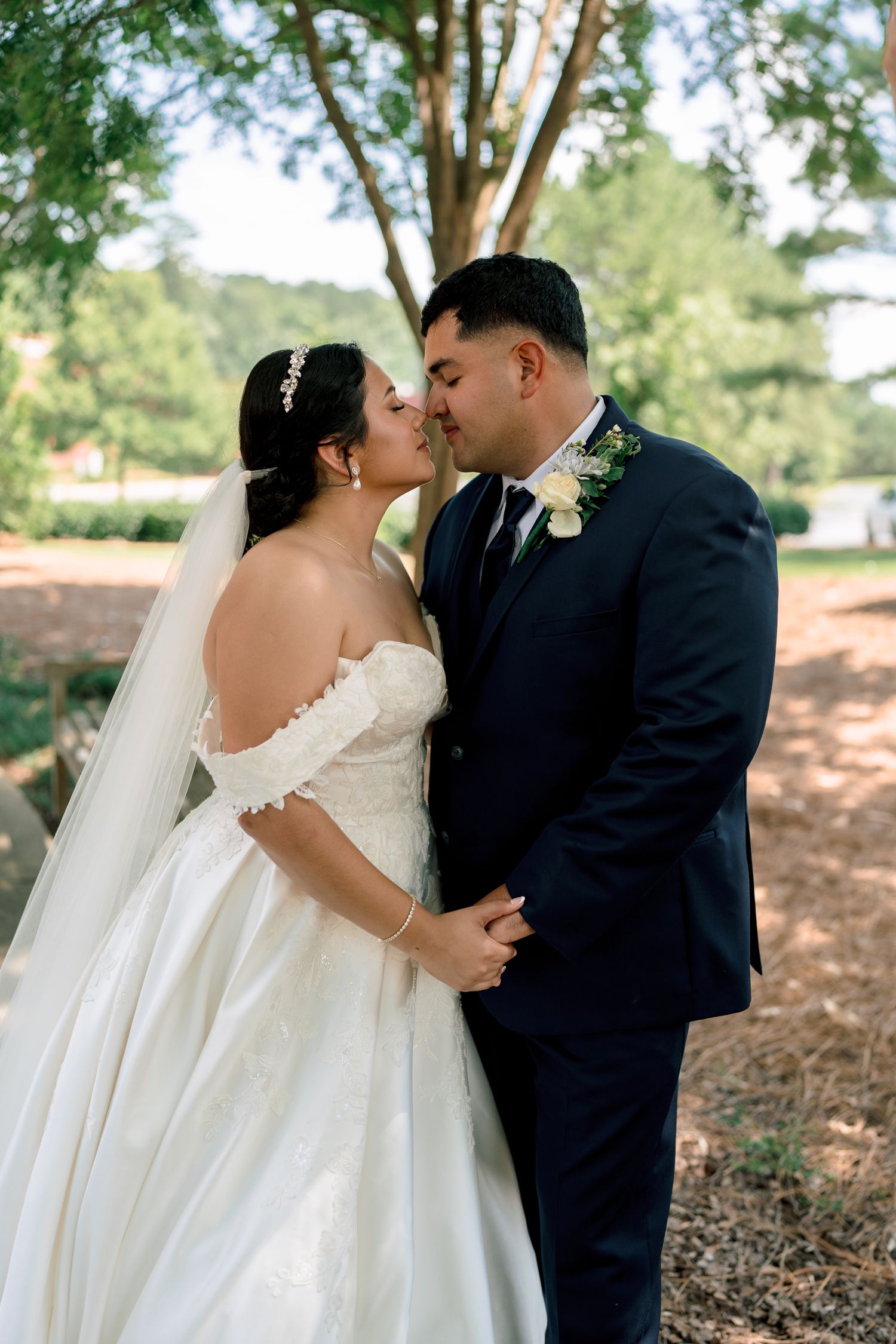 A romantic wedding couple shares a tender kiss outdoors against a backdrop of lush greenery and trees.