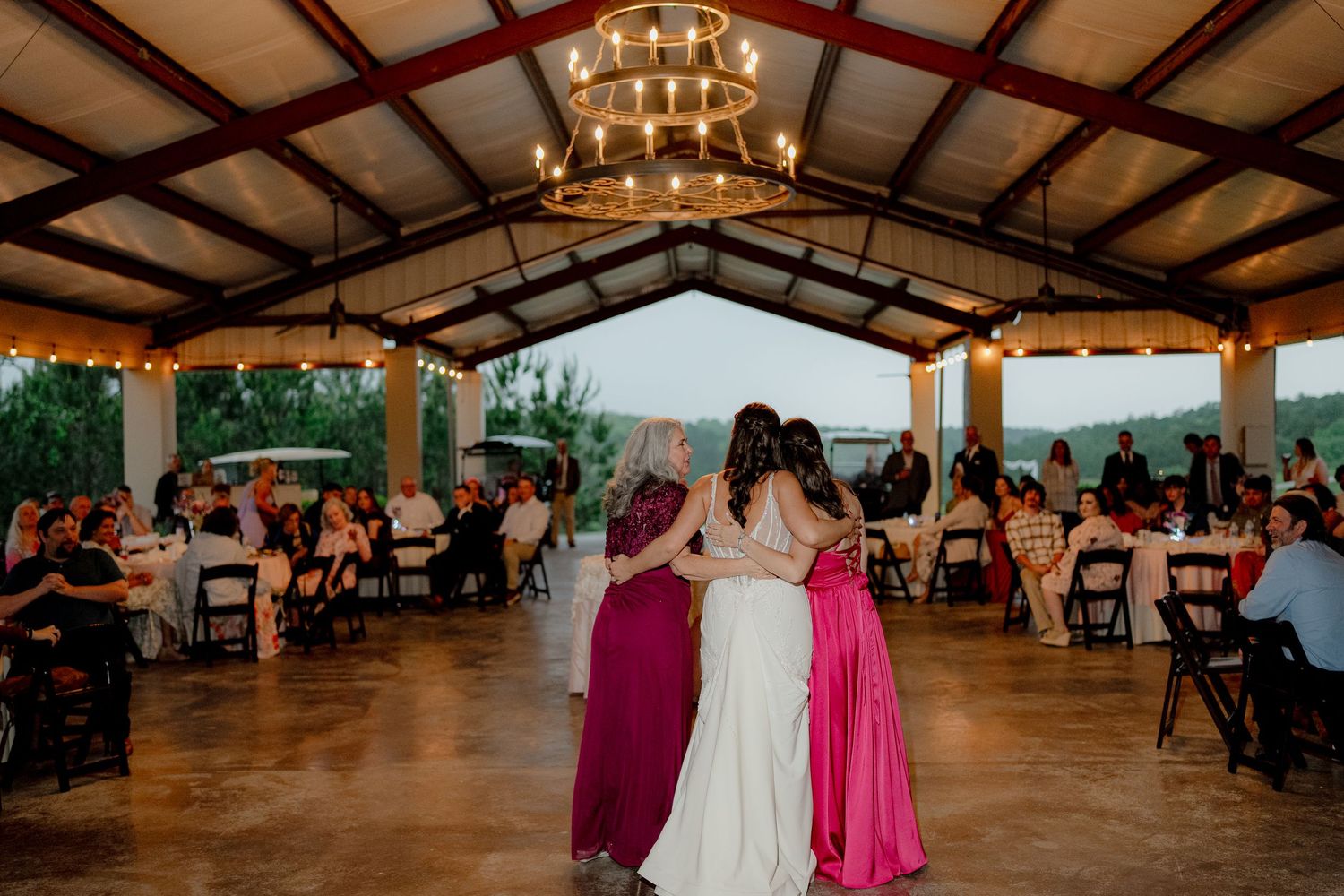 Bride and bridesmaids share a group dance under chandelier lights at an open-air wedding reception.