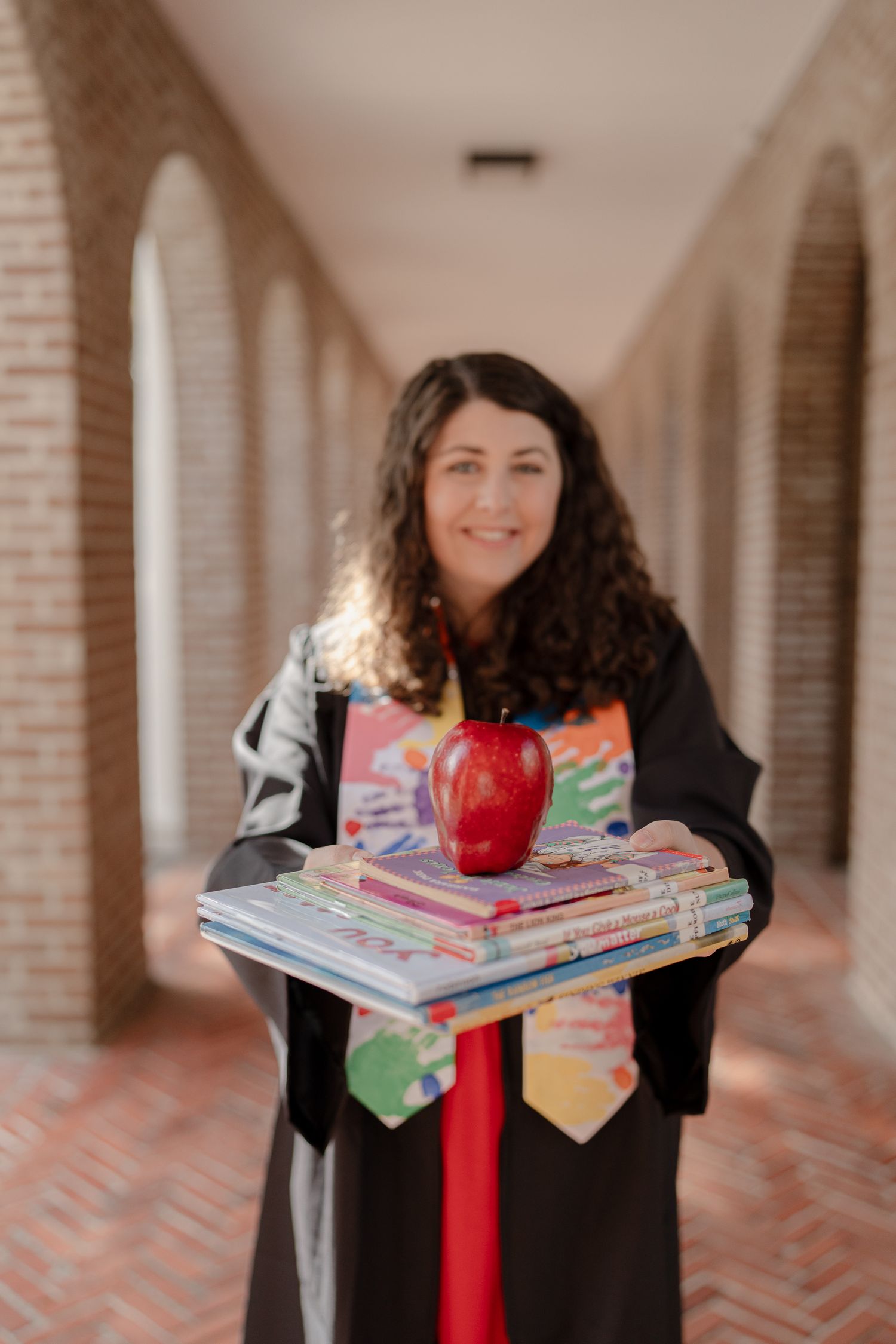 Graduate in black cap and gown holds colorful children's books and a red apple in an arched brick hallway.