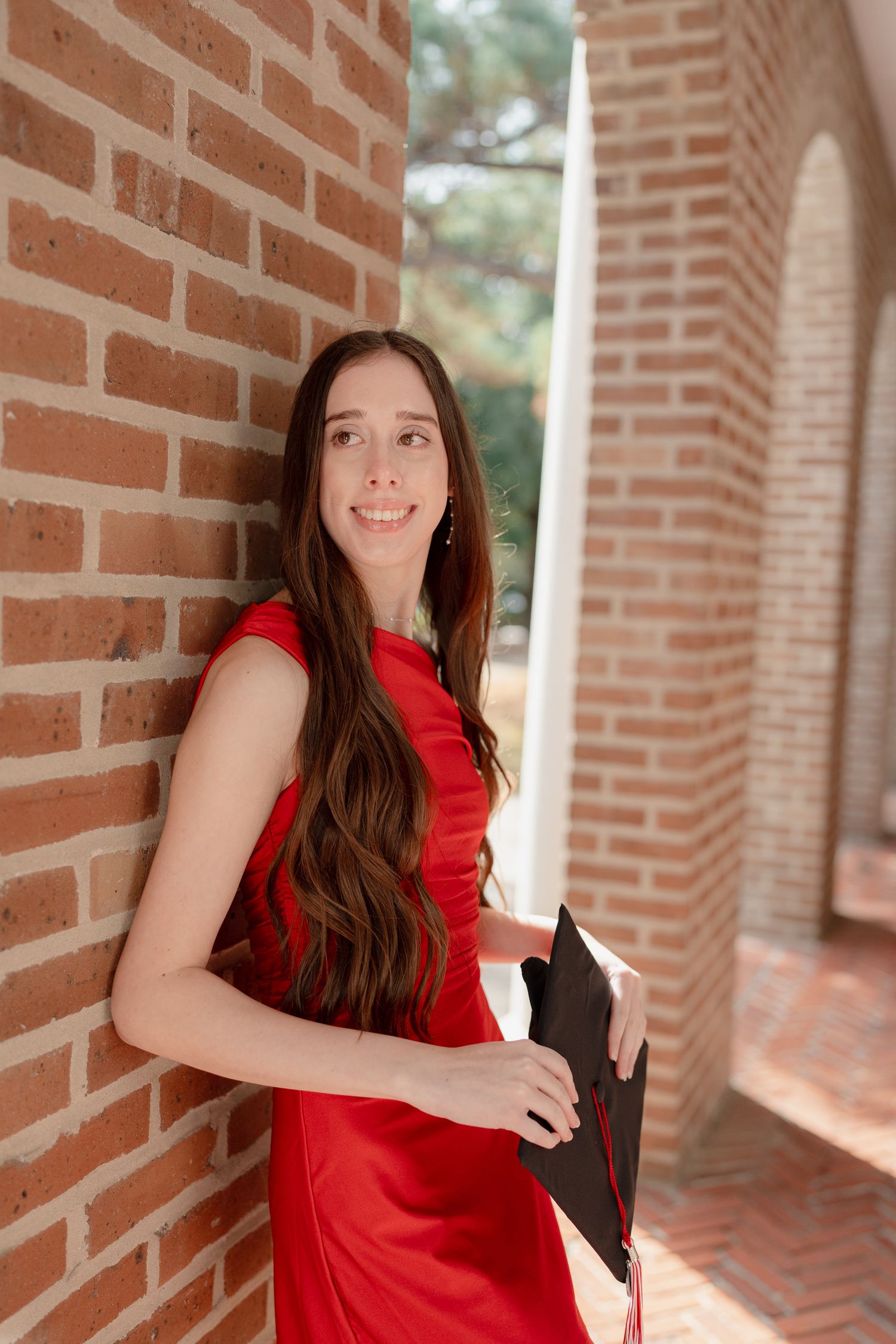 A person in a bright red dress poses against a brick wall with columns in an outdoor setting.