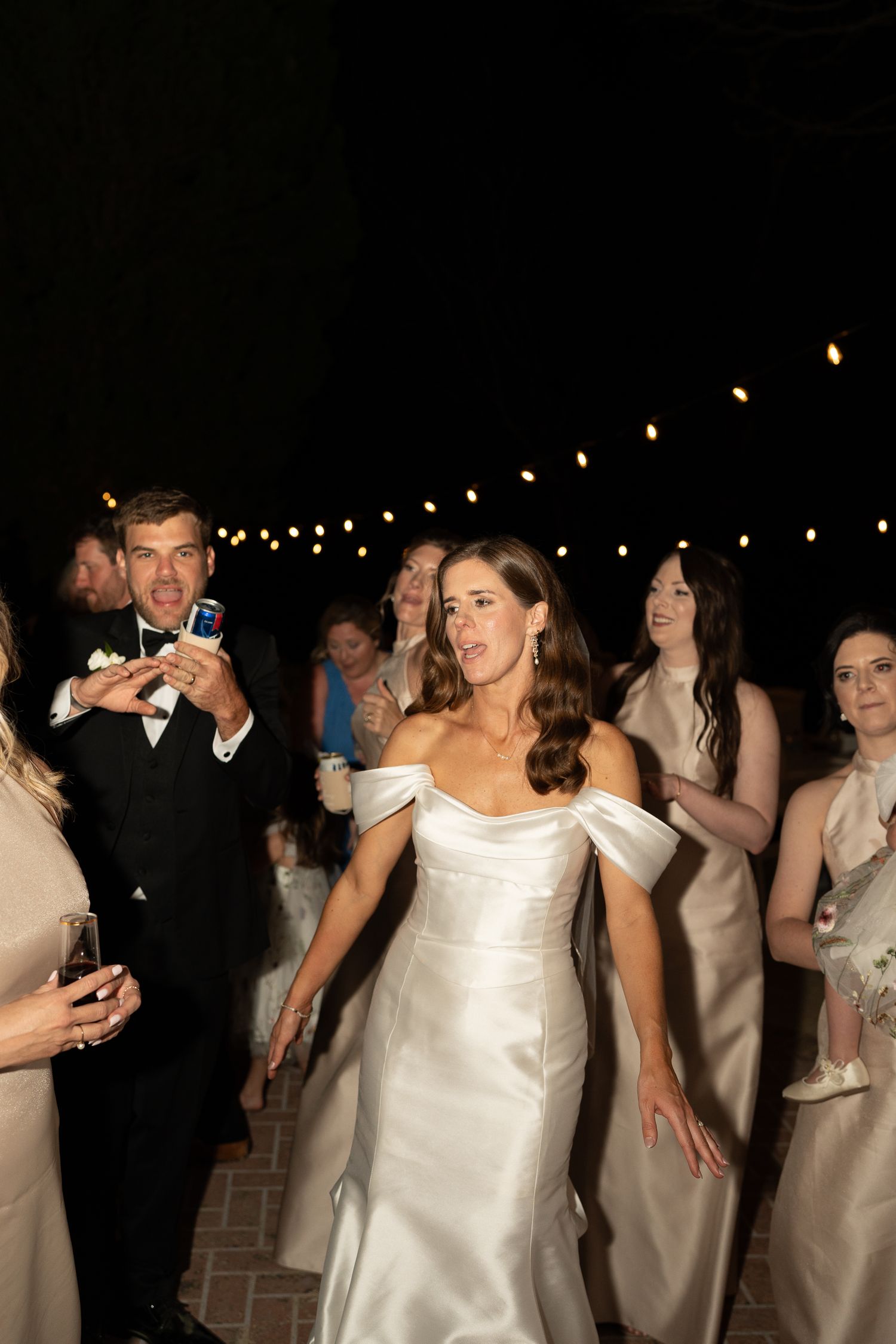 Bridesmaids in white satin dresses dance together under twinkling lights at night.