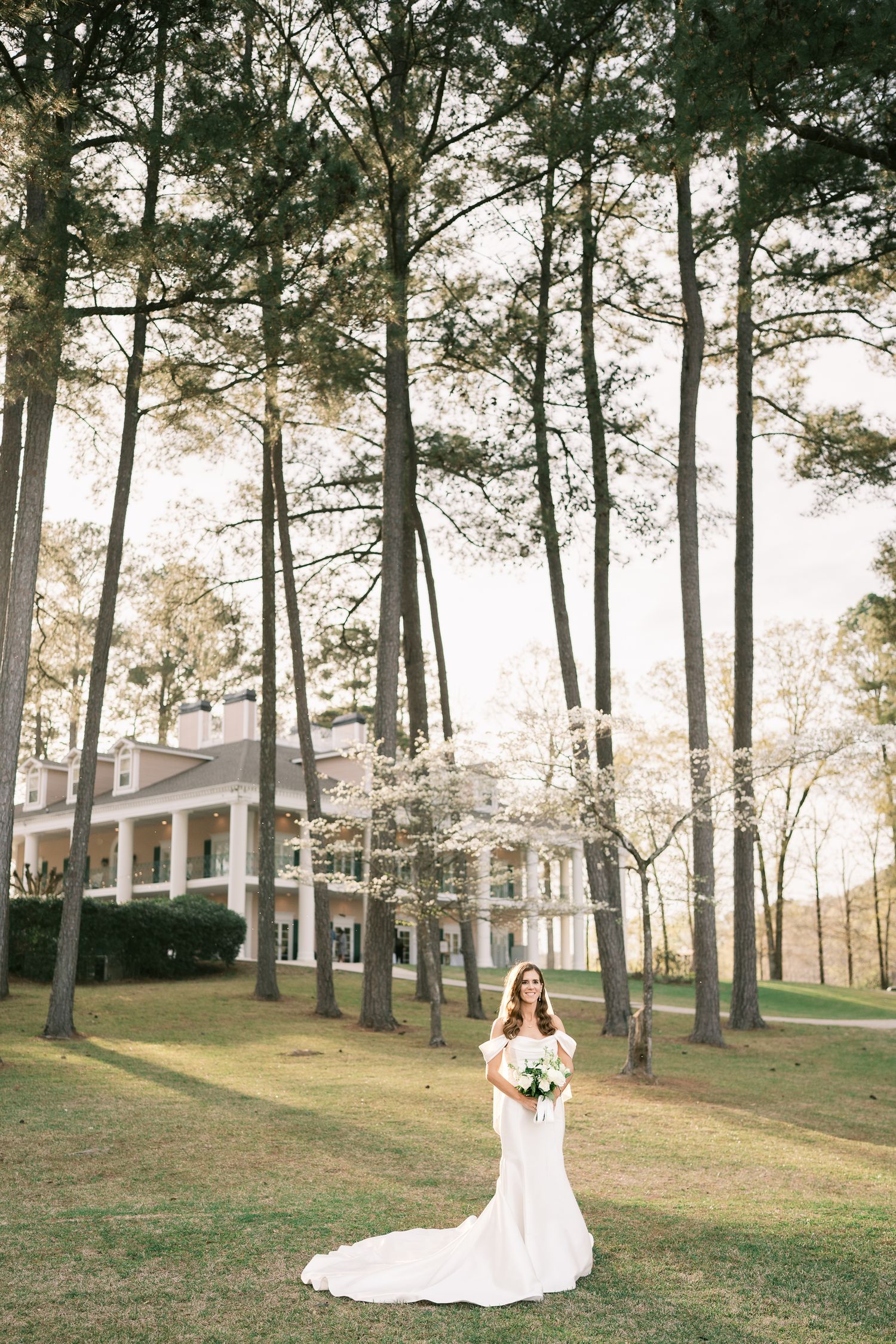 Elegant bridal portrait among tall pine trees with white mansion in background.