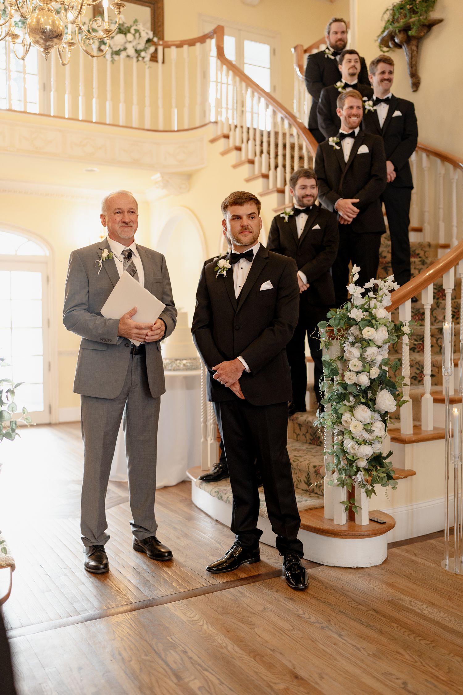 Wedding party arranged on a grand staircase with floral decorations and chandelier.