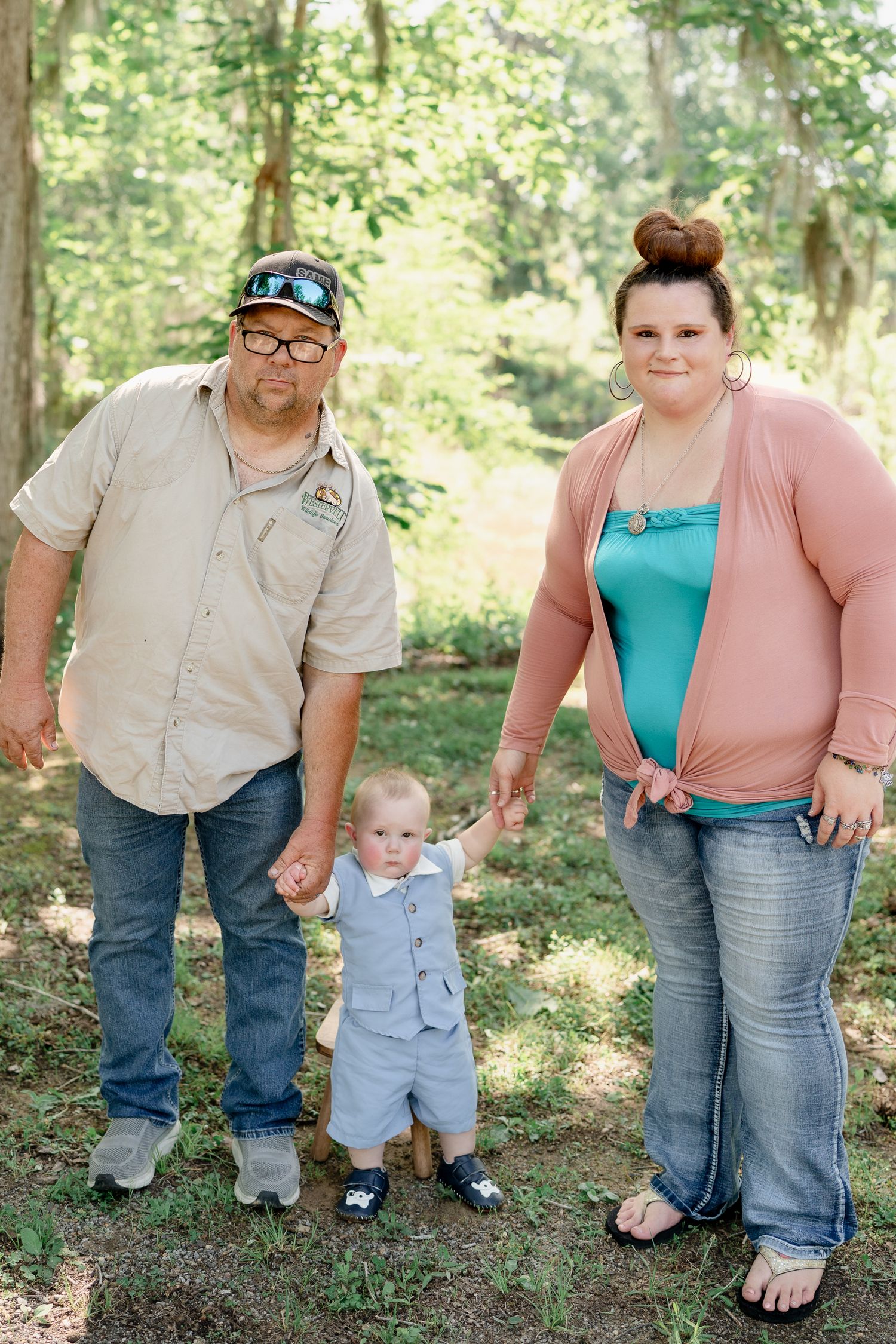 Family of three standing together for a casual portrait in a wooded area.