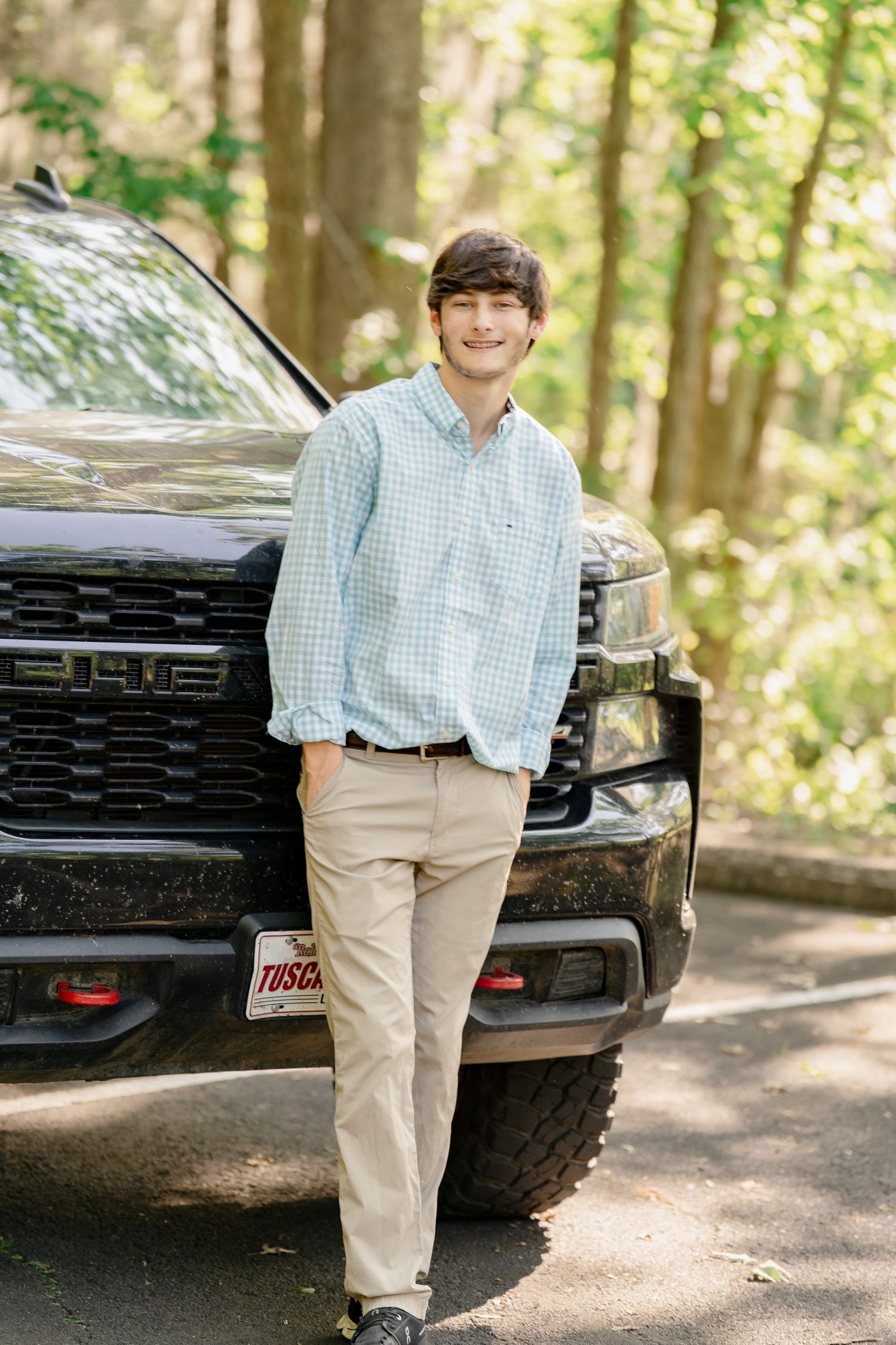 Student in checkered shirt poses with black GMC truck in forest setting.