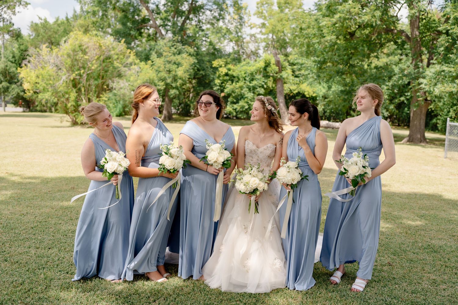 Wedding party in matching light blue bridesmaid dresses holding white floral bouquets pose together outdoors.