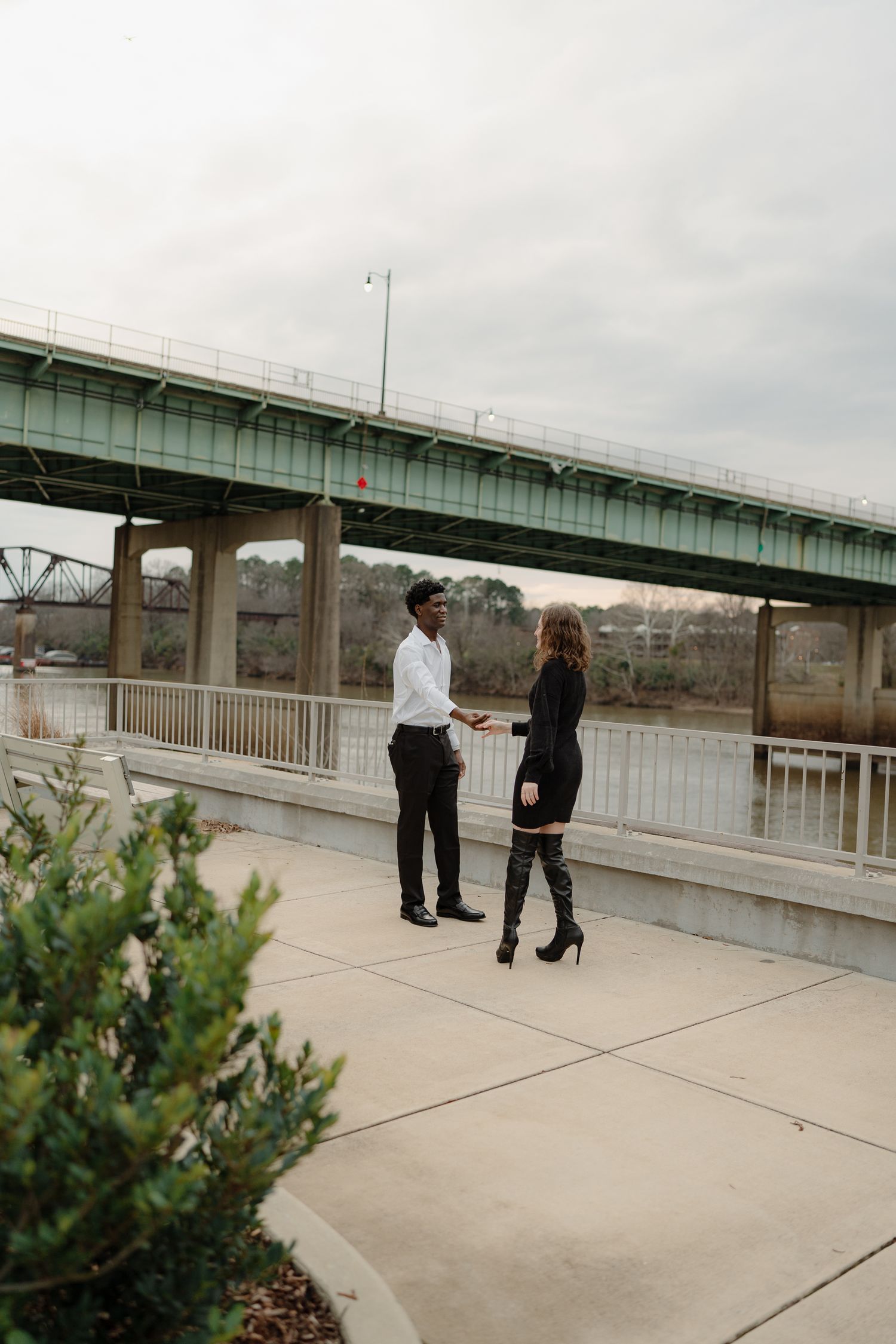Interracial couple talking on a riverside walkway beneath a bridge on an overcast day.