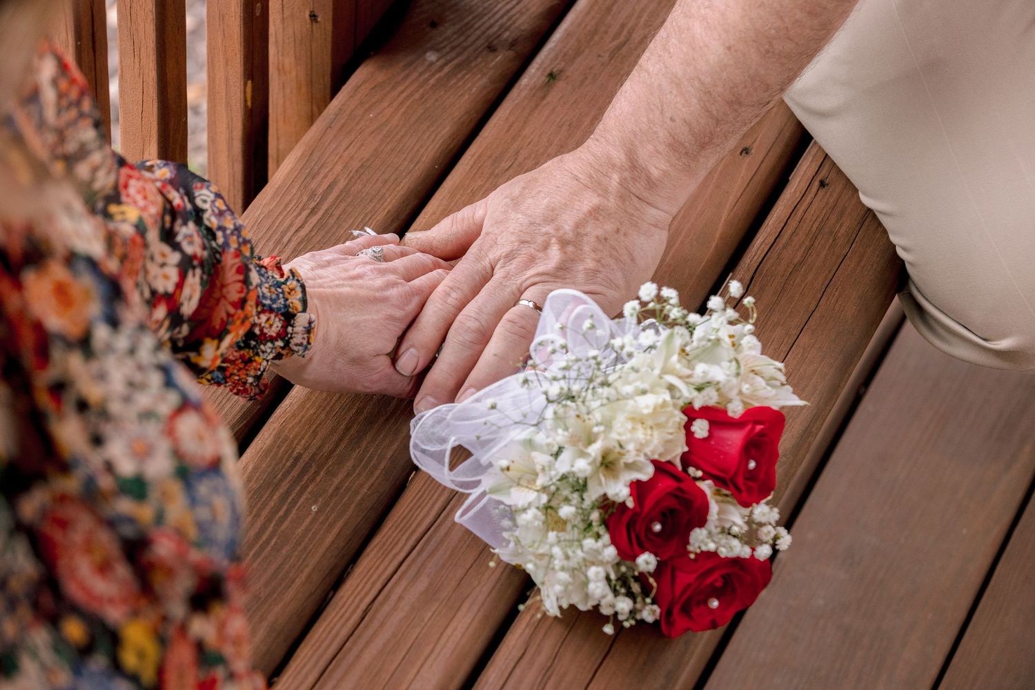 Close-up of couple holding hands on wooden bench with a red and white wrist corsage bouquet.
