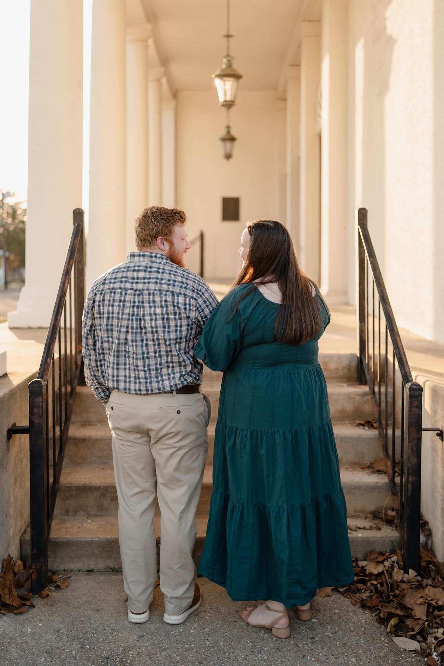 Couple in coordinating outfits walks up historic building steps, woman in teal dress and man in plaid shirt.