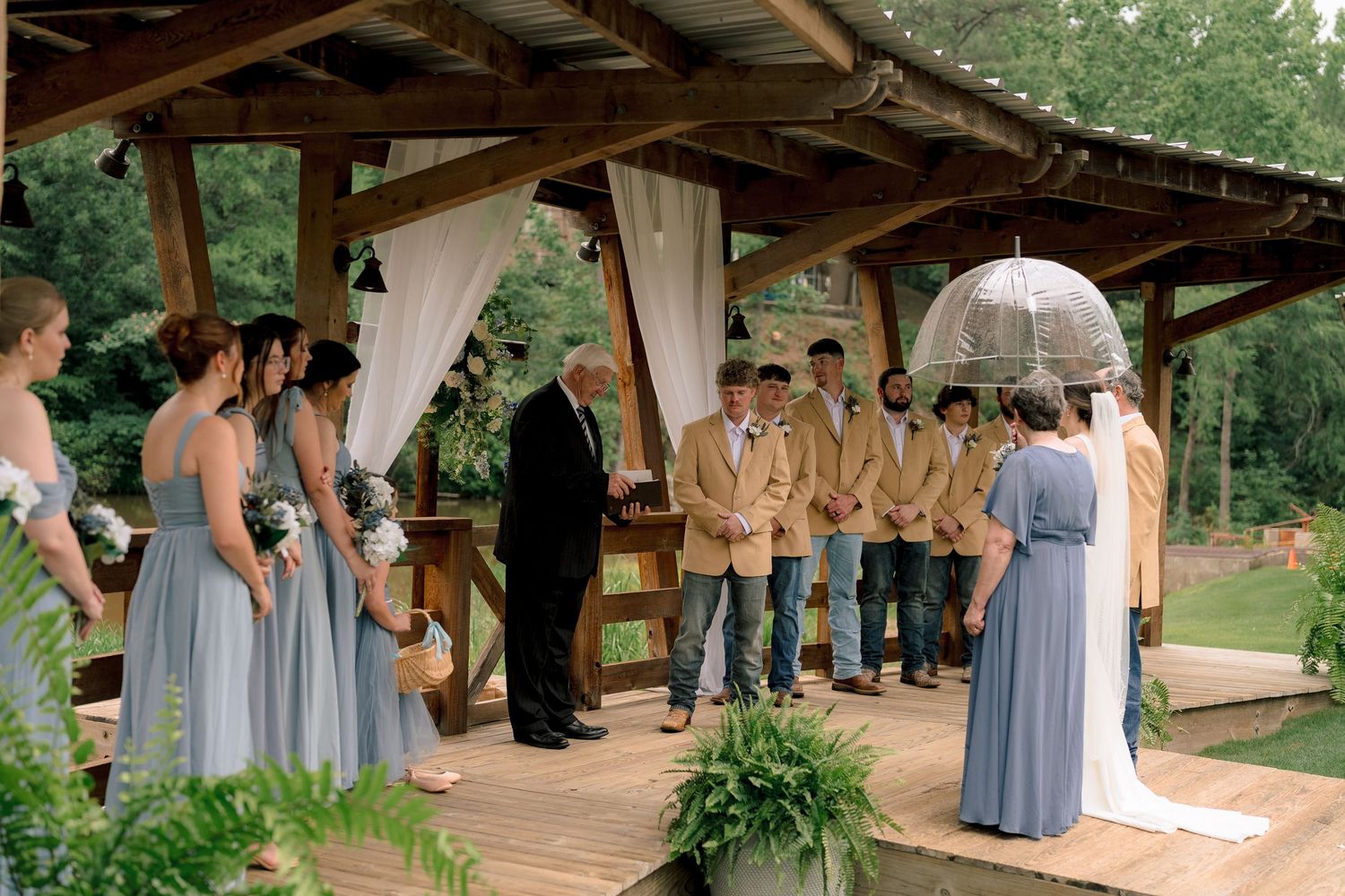 Outdoor rustic wedding ceremony under wooden pavilion with bridesmaids in blue and groomsmen in tan.