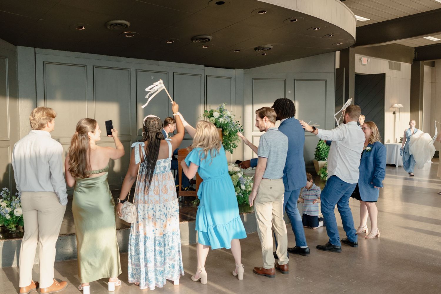 A panoramic view shows wedding guests lined up in a row inside a bright indoor venue.