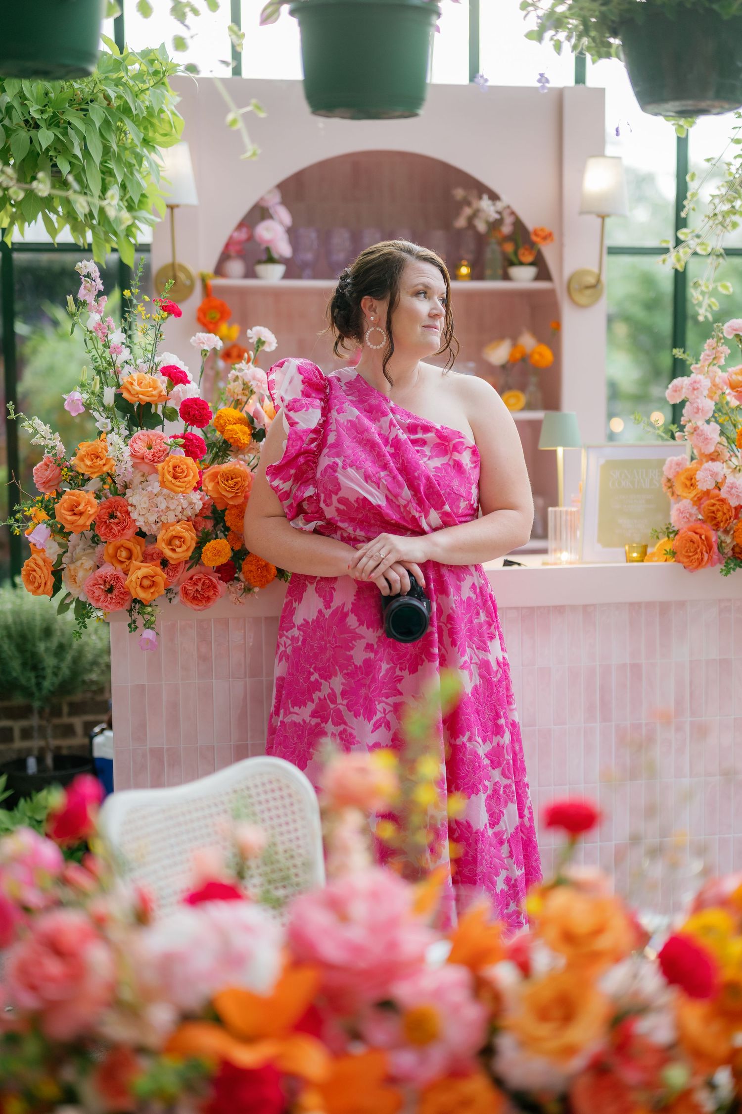 Woman in pink floral one-shoulder dress surrounded by colorful flower arrangements in a bright, plant-filled venue.
