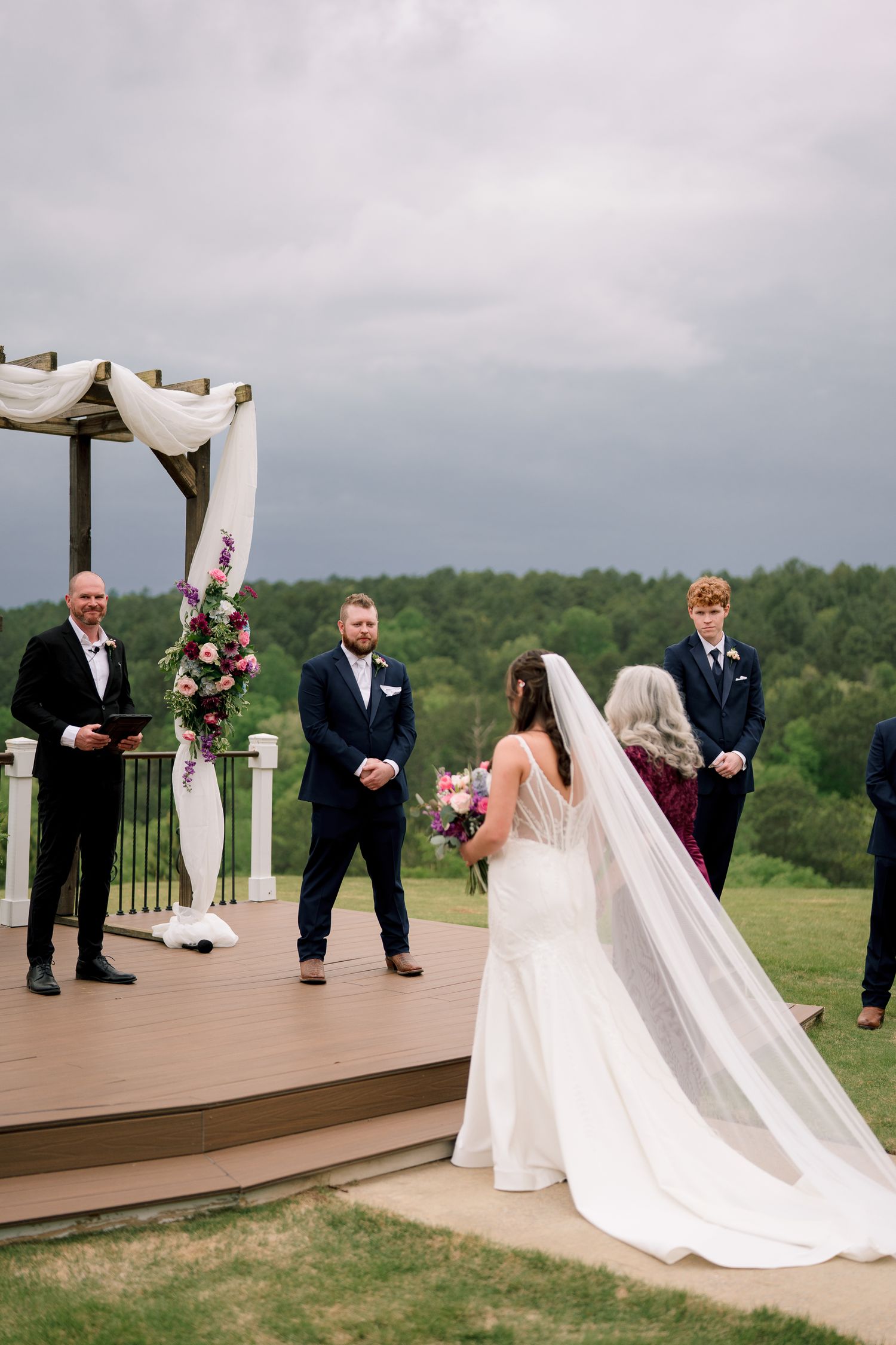 Bride walks toward groom at outdoor wedding ceremony altar decorated with white draping and flowers.
