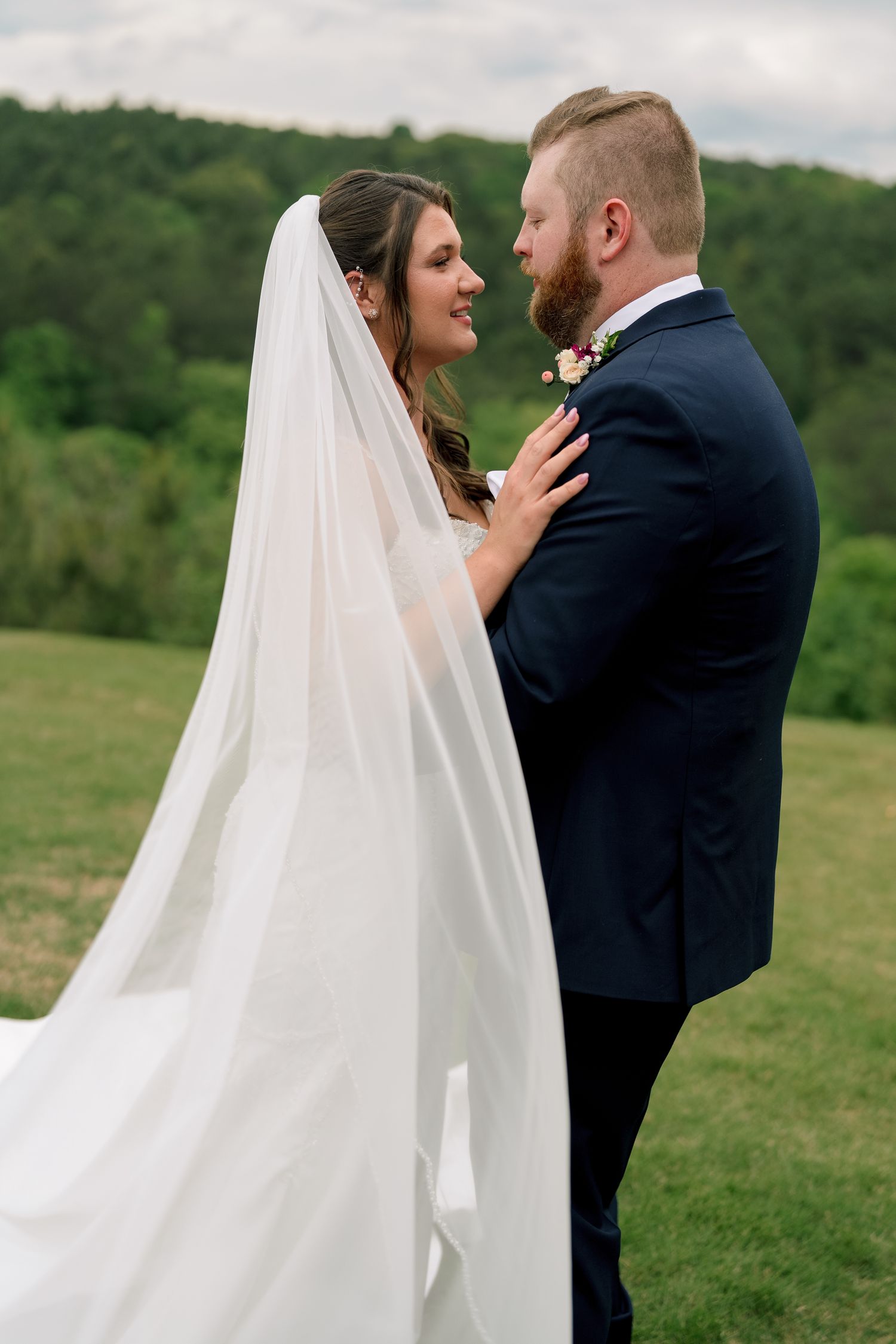 Bride and groom sharing romantic moment outdoors, her flowing veil trailing as she touches his navy suit.