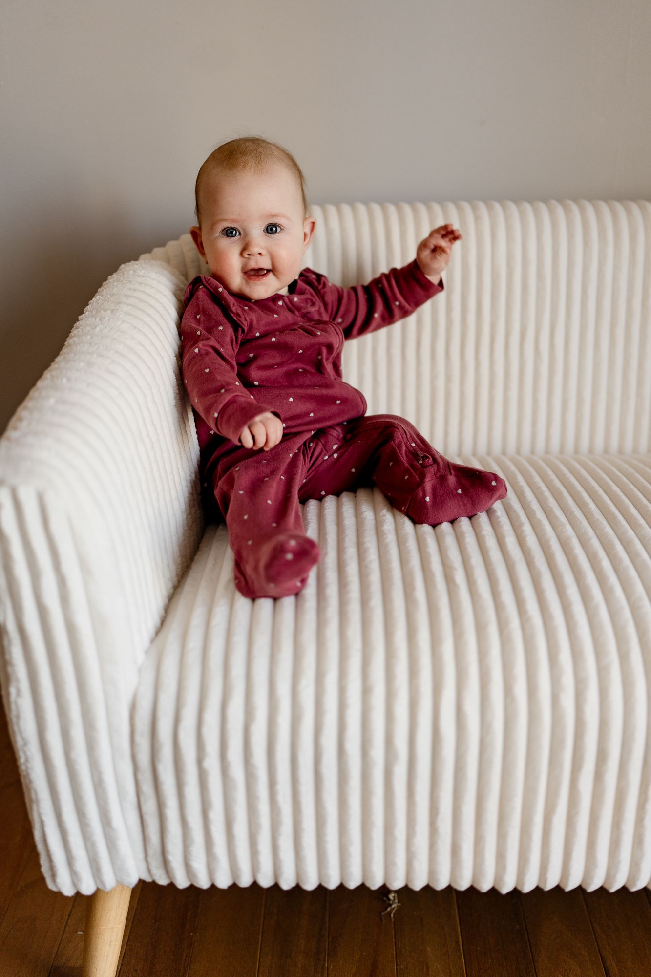 Baby in burgundy outfit smiles and gestures while sitting on white textured couch.