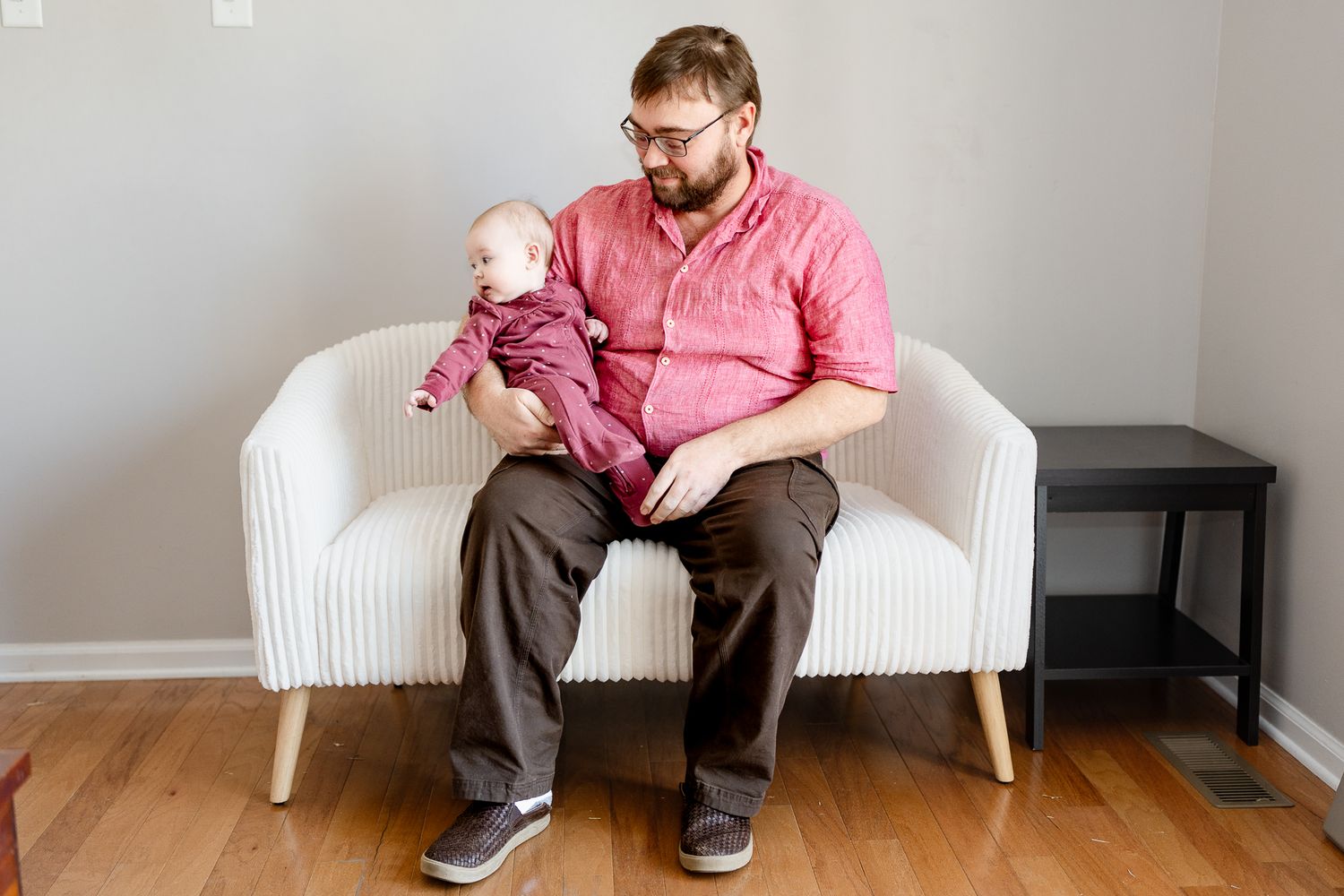 Adult and baby sit together on white modern loveseat in minimalist room setting.