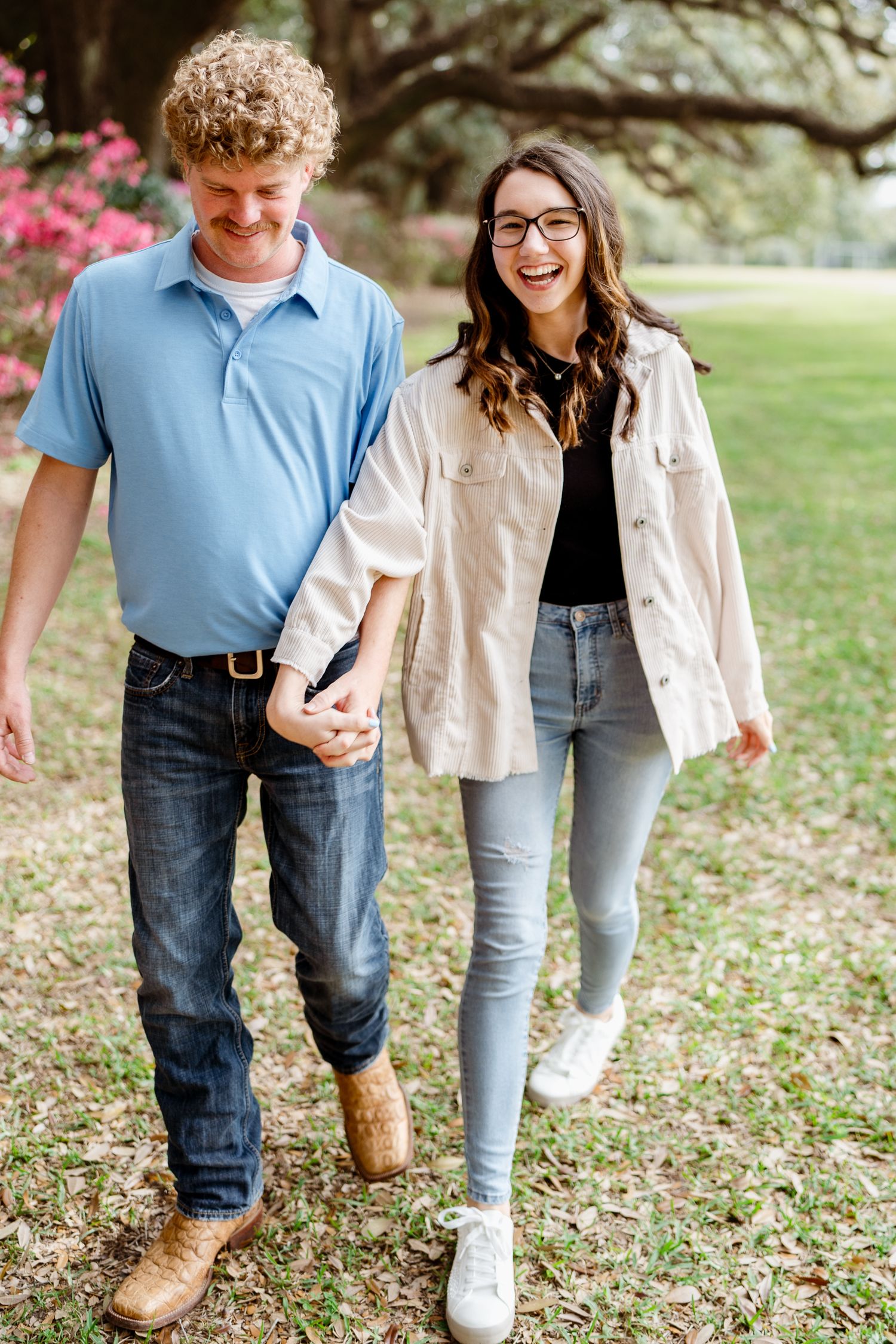 A couple in casual attire walking together under a canopy of oak trees with scattered flowers in the background.