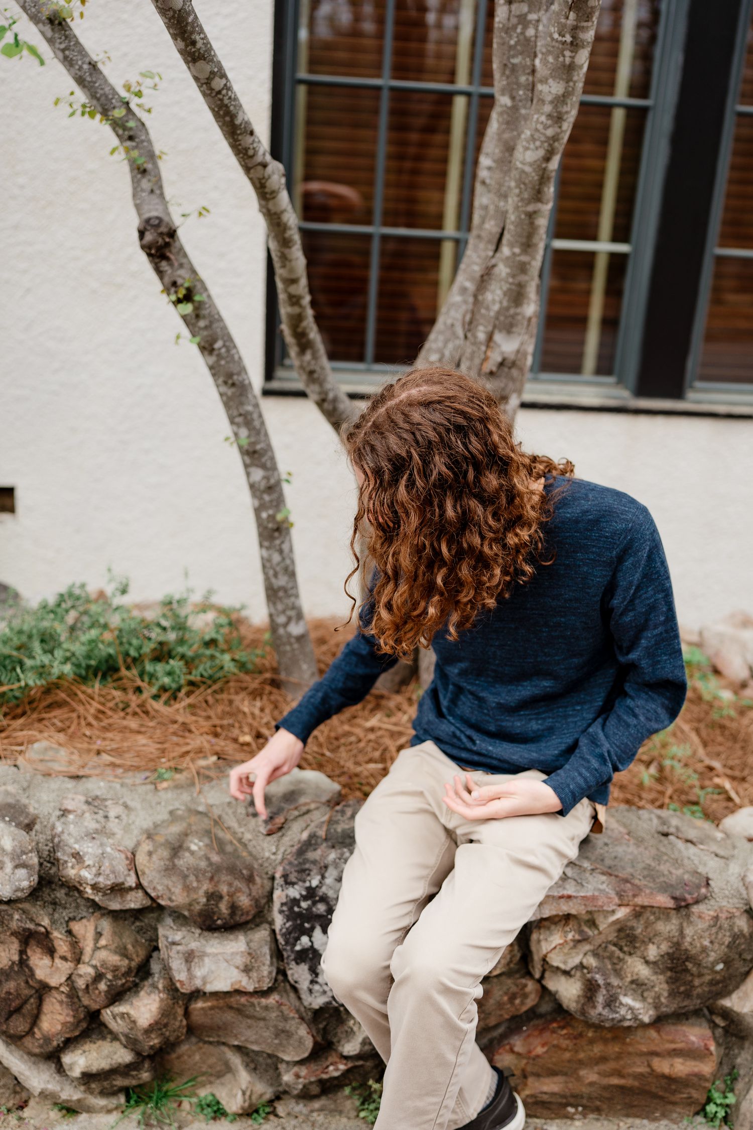 Person with curly hair sitting on stone wall near building with white exterior.
