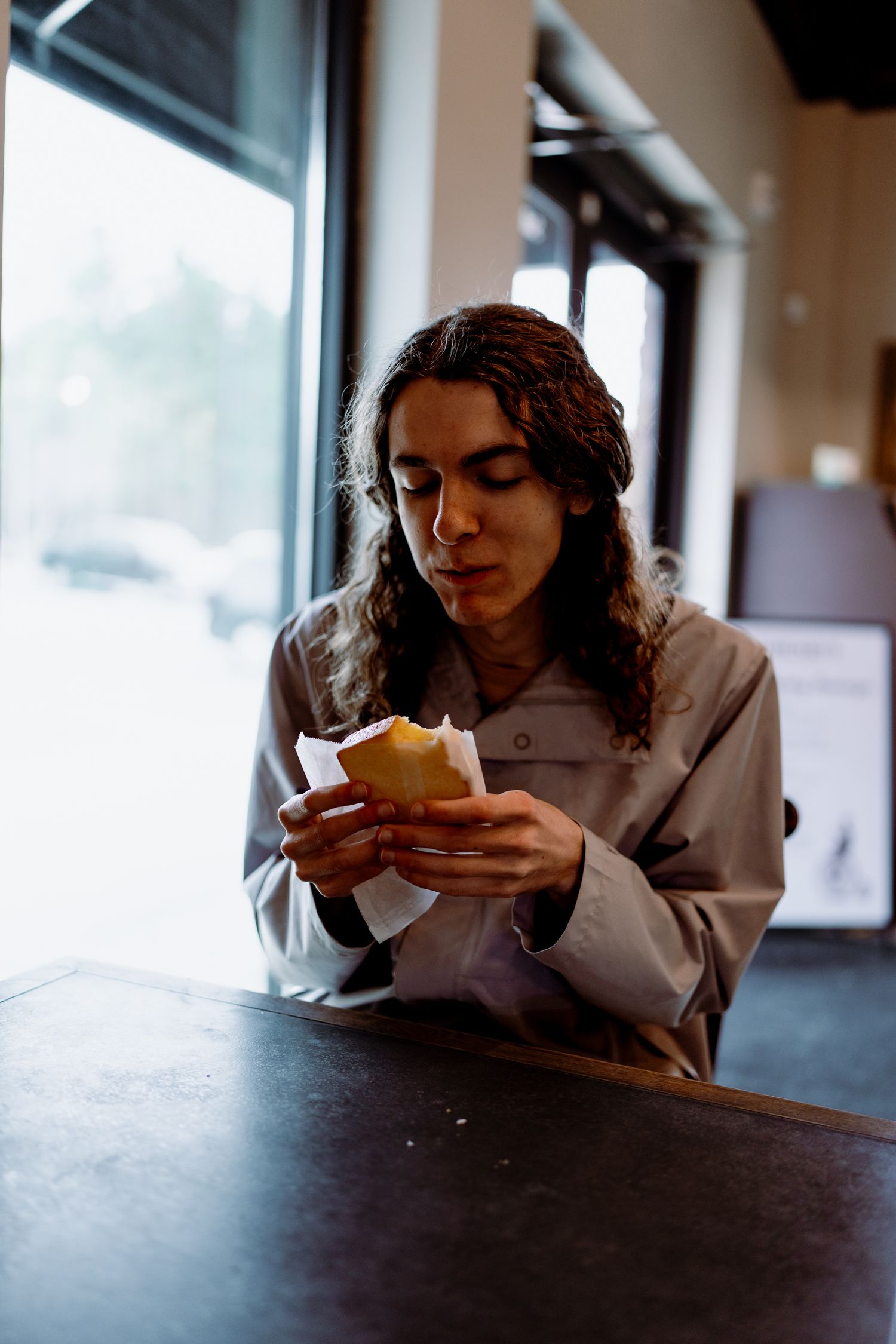 Person in casual attire eating food while seated at dark wooden table near window.