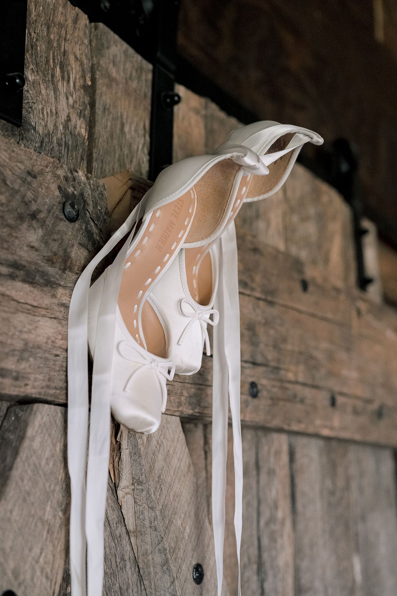 White bridal ballet flats with ribbon ties hanging on a rustic wooden barn door.