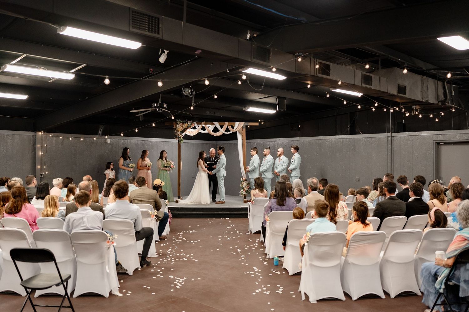 An indoor wedding ceremony with white chairs and guests seated in rows facing a stage with string lights overhead.