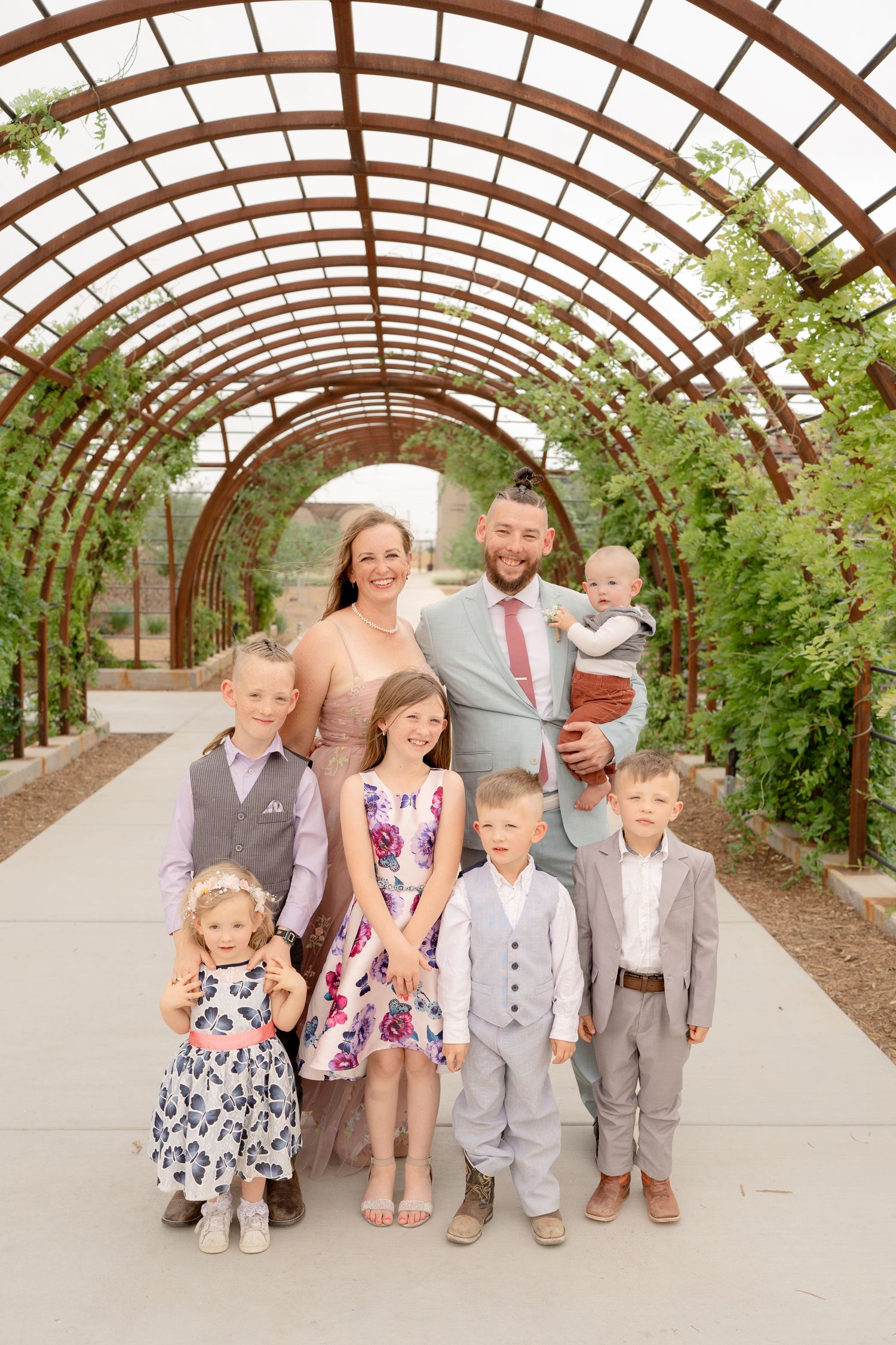 Family group in coordinated outfits sits together under a curved garden arbor walkway.