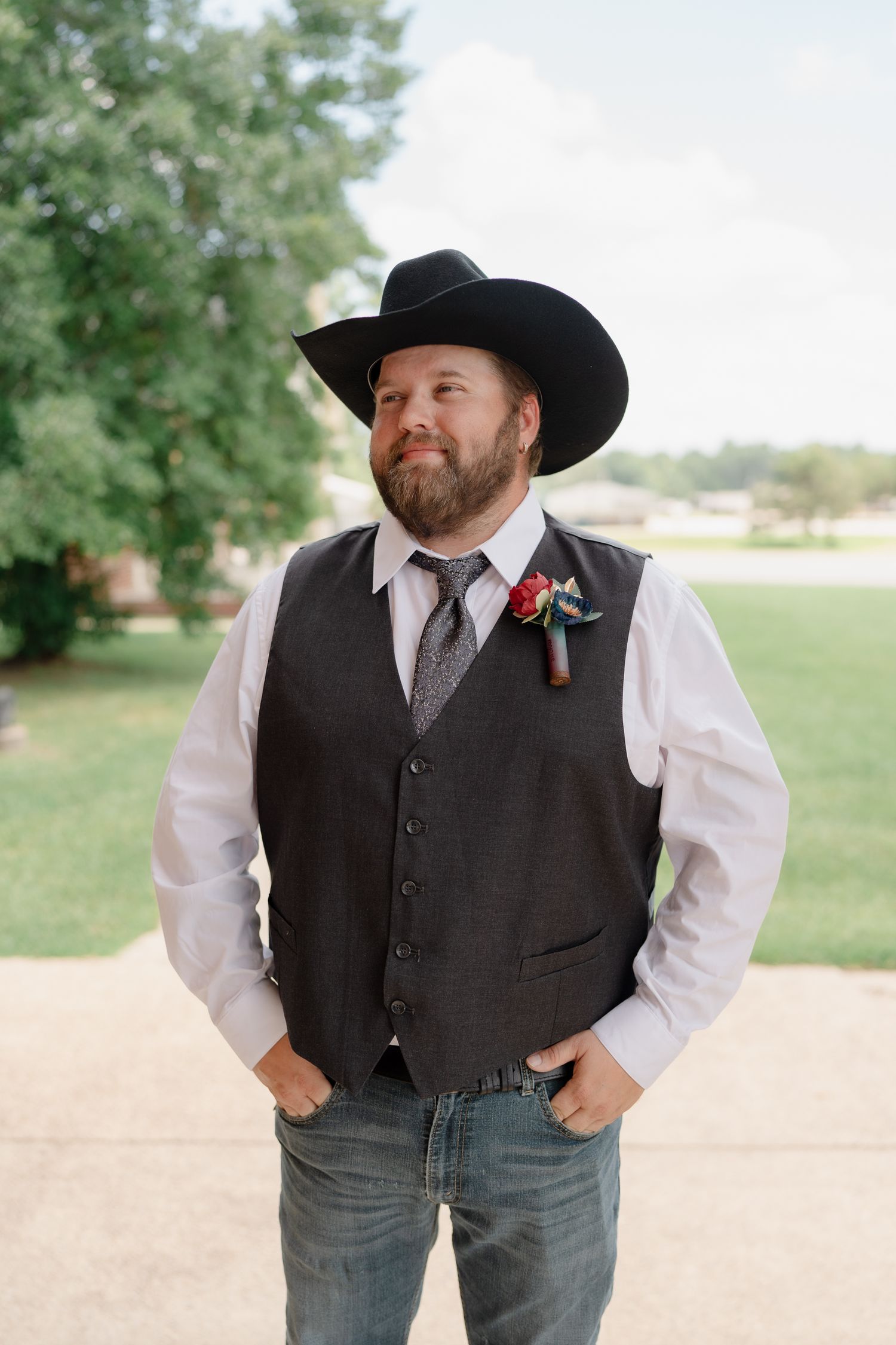 A western-styled figure in a black cowboy hat, dark vest and white shirt poses casually outdoors against a natural backdrop.
