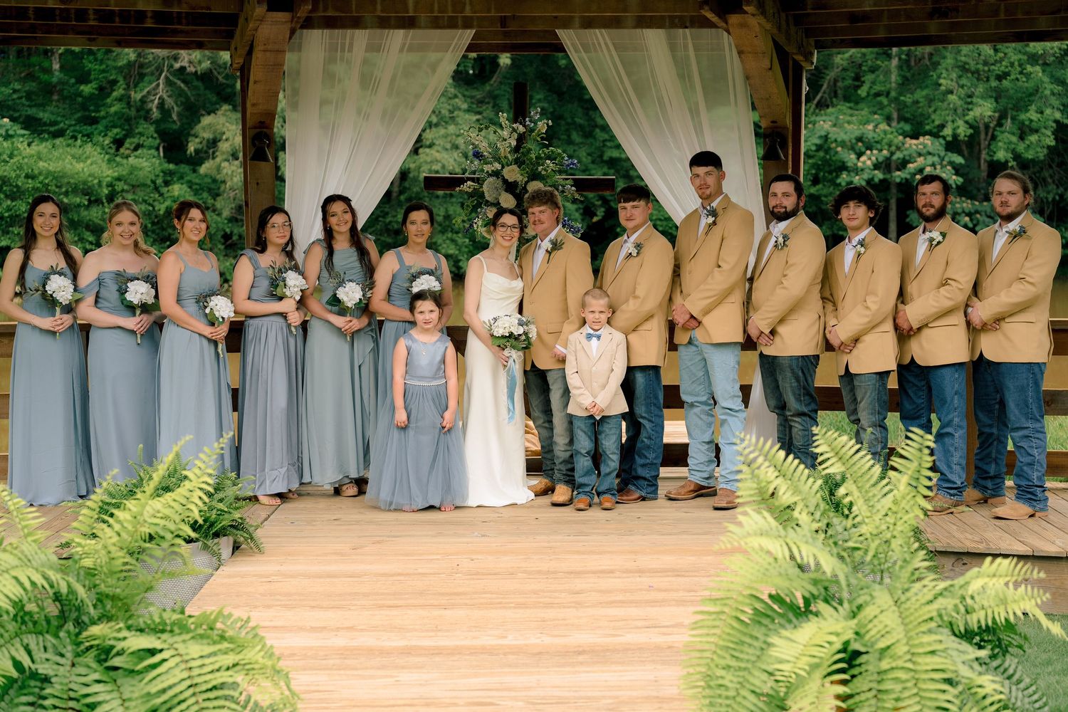 Wedding party poses under rustic wooden pavilion with bridesmaids in gray gowns and groomsmen in tan jackets.