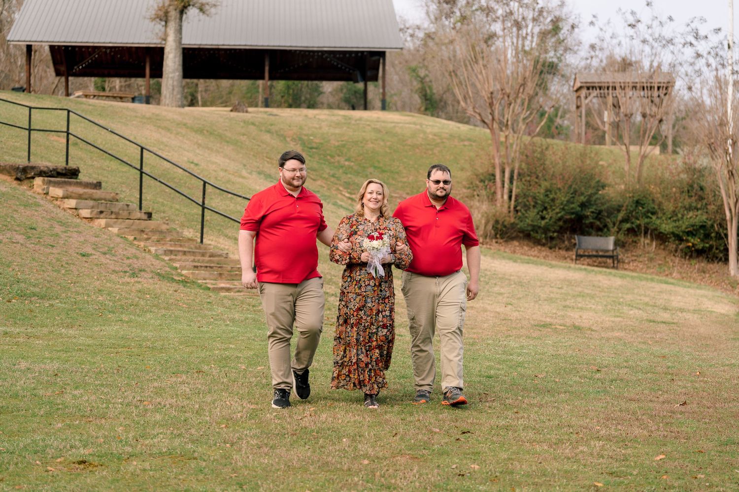 A woman in a floral dress walks outdoors with two men in red shirts on a green lawn.