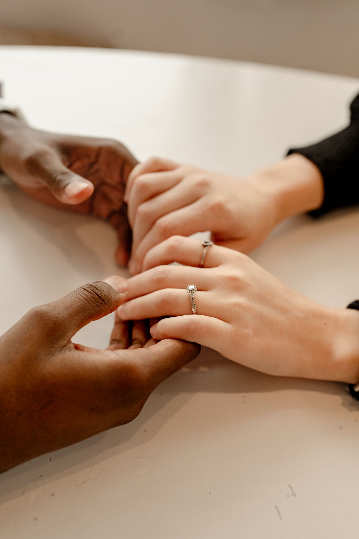 Close-up of interracial couple holding hands on white table, woman wearing silver rings on her fingers.