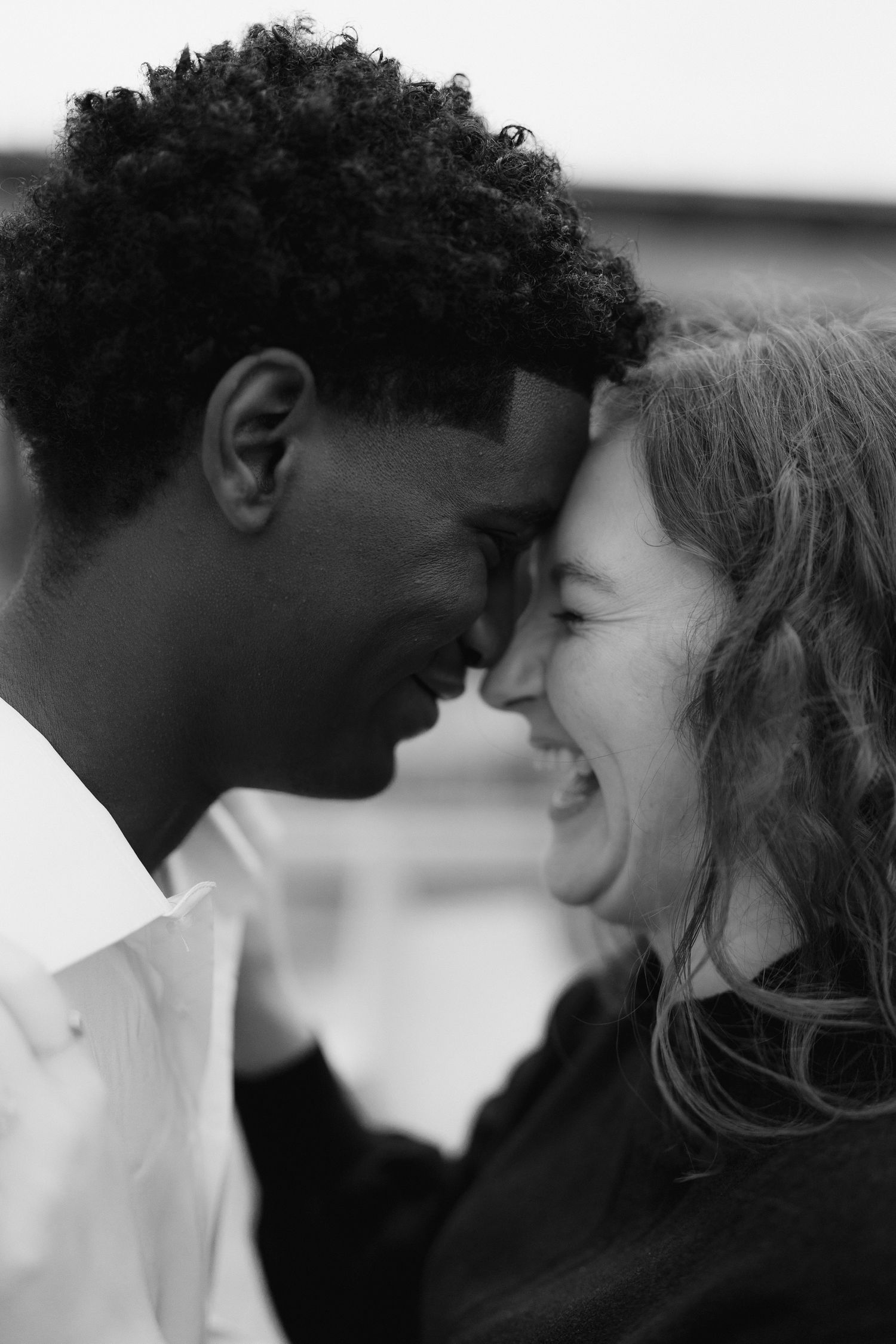 A joyful interracial couple laughs together with foreheads touching in a tender black and white portrait.