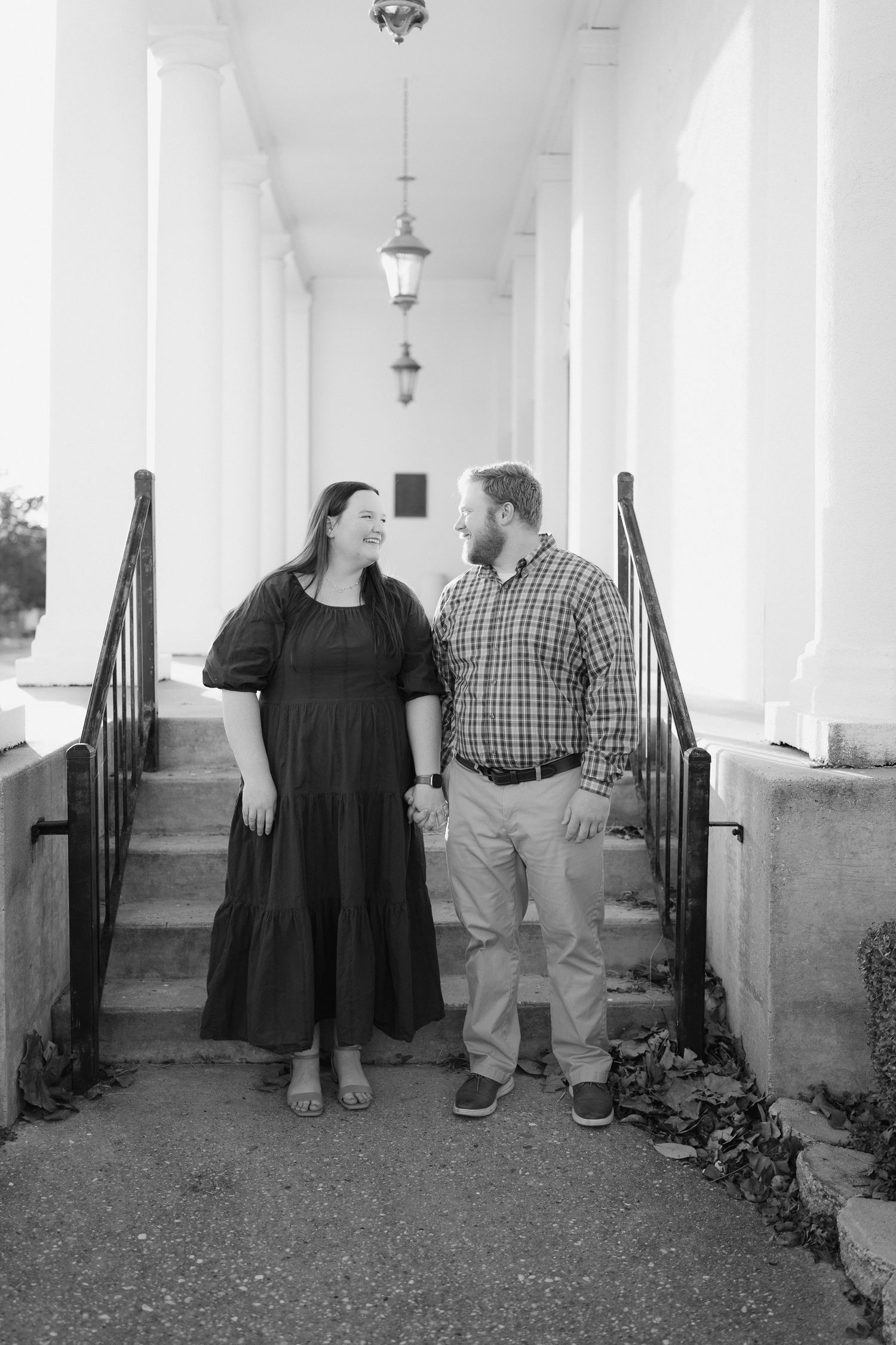 Couple holding hands and smiling at each other beside white columns and steps in black and white.