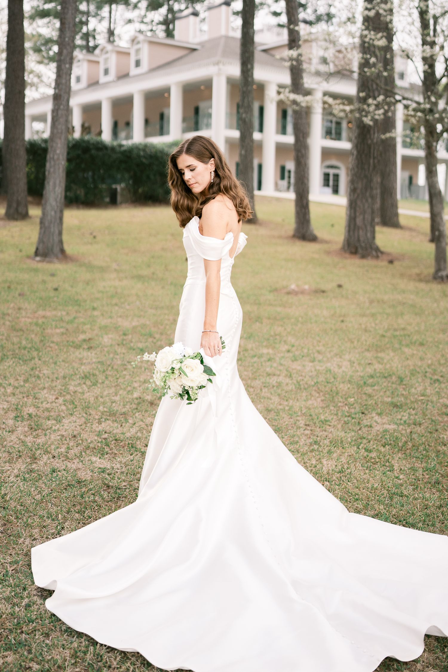 Bride in off-shoulder gown with cathedral train stands before Southern colonial mansion.